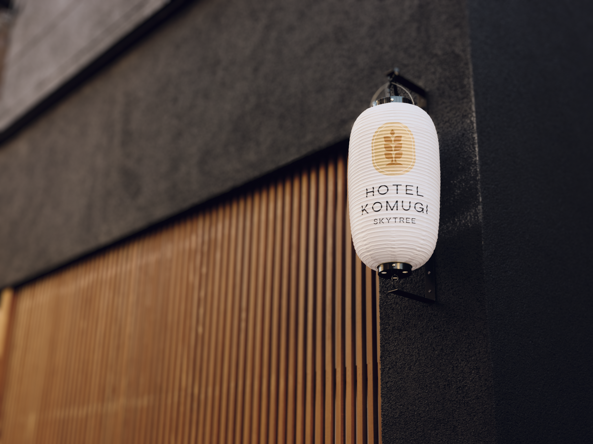 A white paper lantern with black text and a beige logo hanging on a black wall outside a building. The text on the lantern reads 'HOTEL KOMUGI SKYTREE'.
