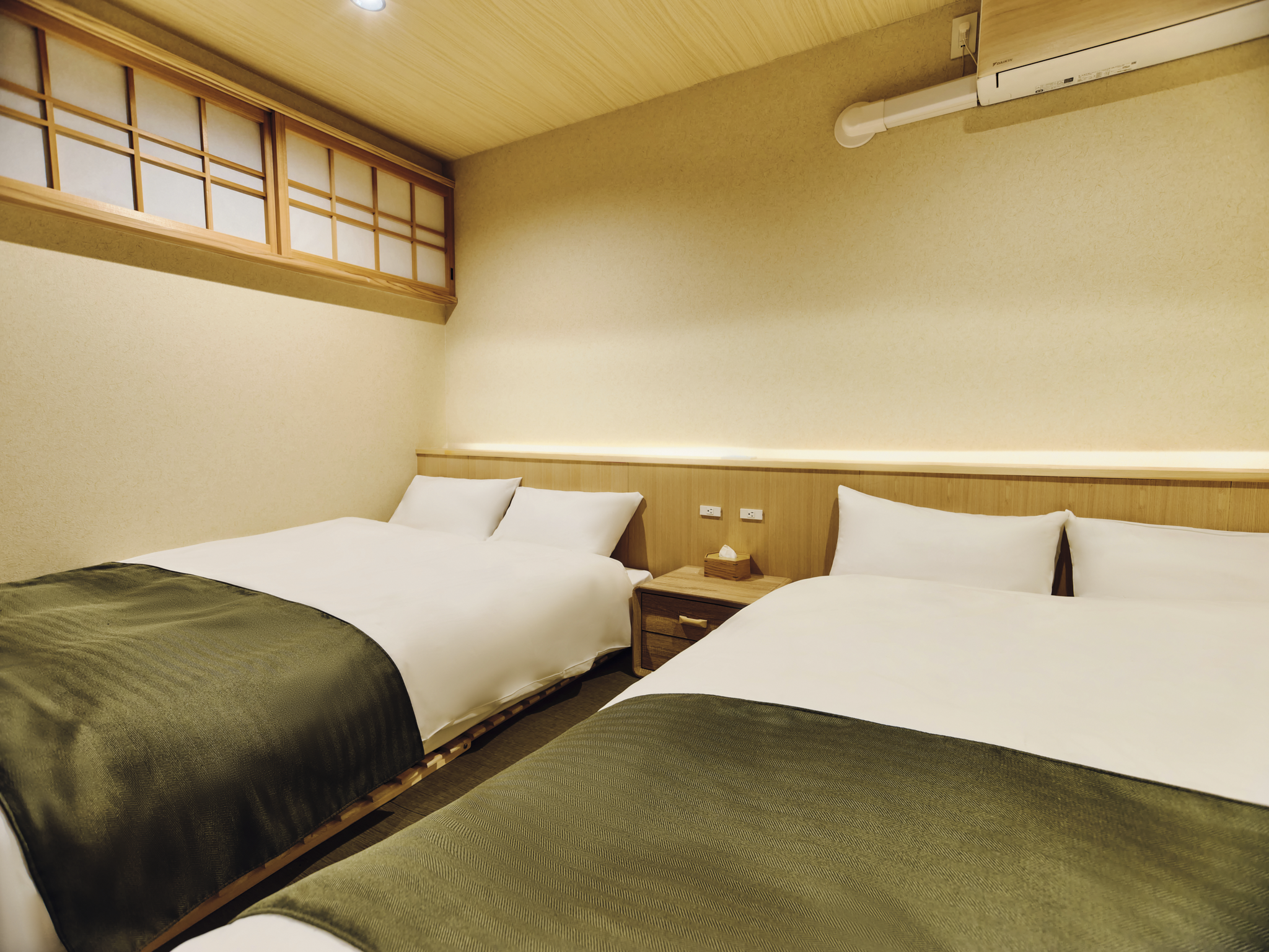 A hotel room with two beds, each with white bedding and green bed runners, a wooden nightstand with a tissue box, beige walls, a wooden ceiling panel, and an air conditioning unit.