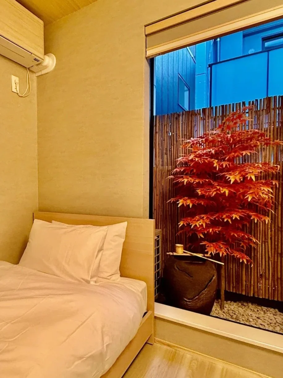 A minimalist bedroom with a bed, white bedding, and pillows. There's a large window showing an outdoor Japanese maple tree with red leaves, a bamboo privacy screen, and a stone garden. An air conditioning unit is mounted on the wall.
