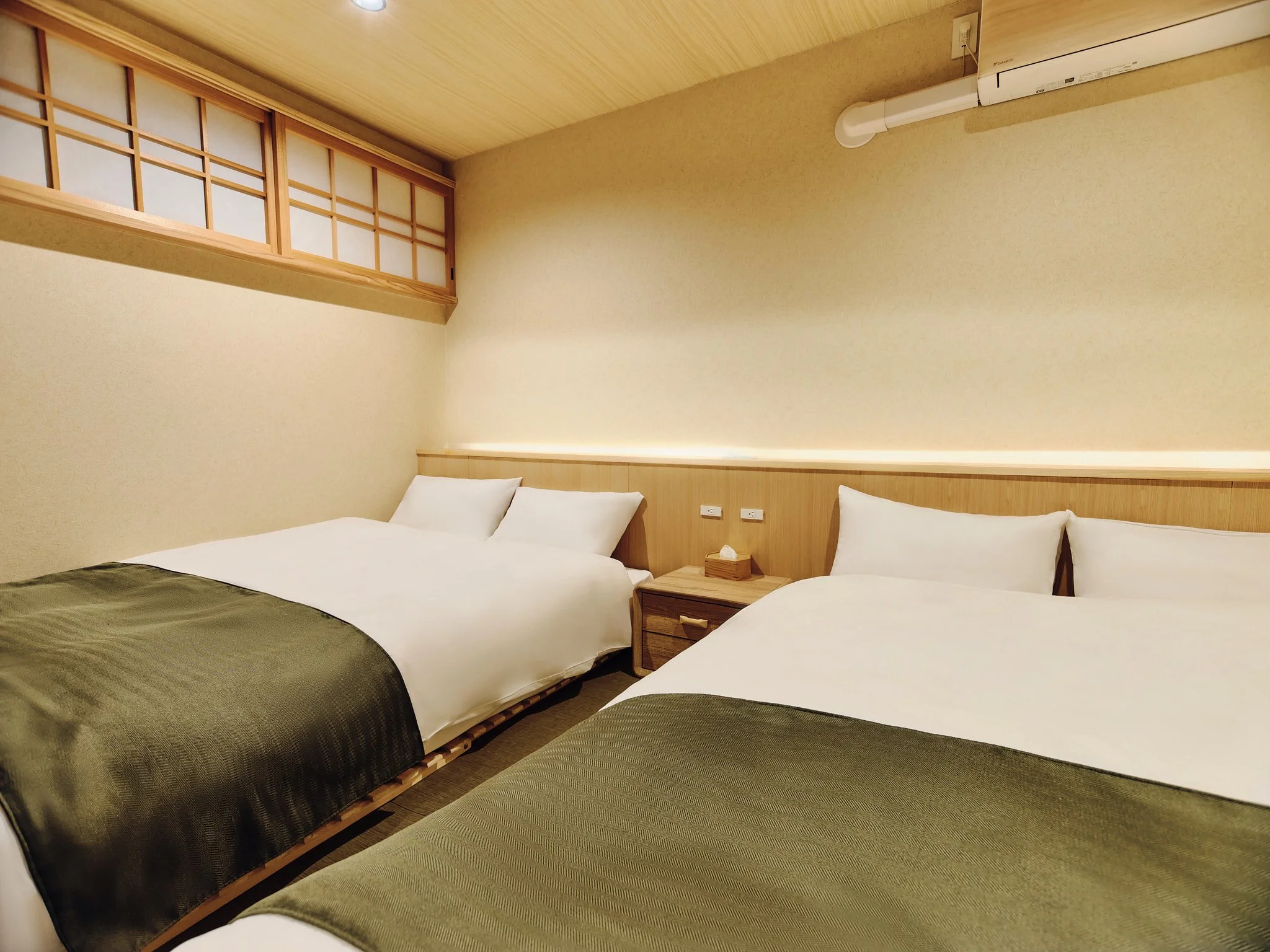 Two neatly made beds with white sheets and green bed runners in a minimalist hotel room with beige walls, wooden accents, and an air conditioning unit.