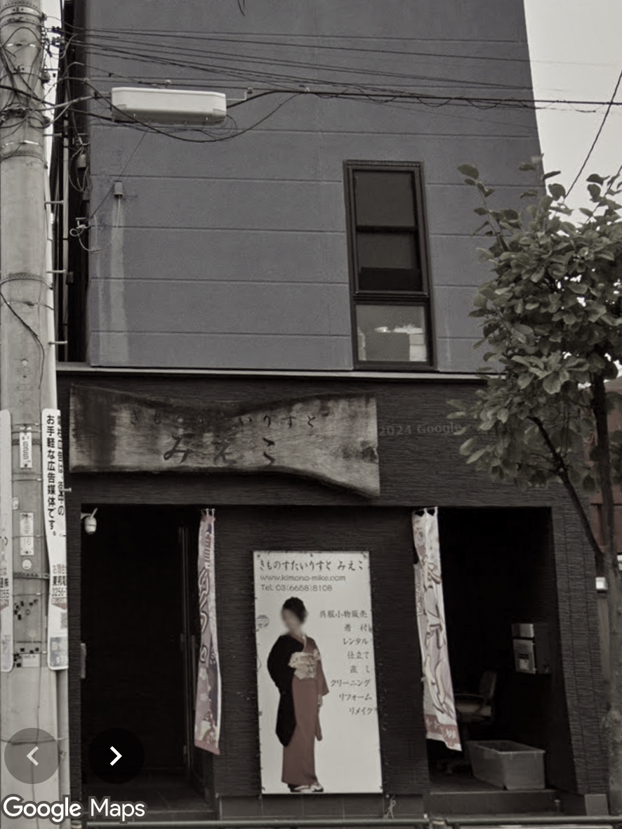 A storefront with a wooden sign, a woman in traditional kimono, and promotional banners outside. The building has a gray upper facade and a dark lower section.