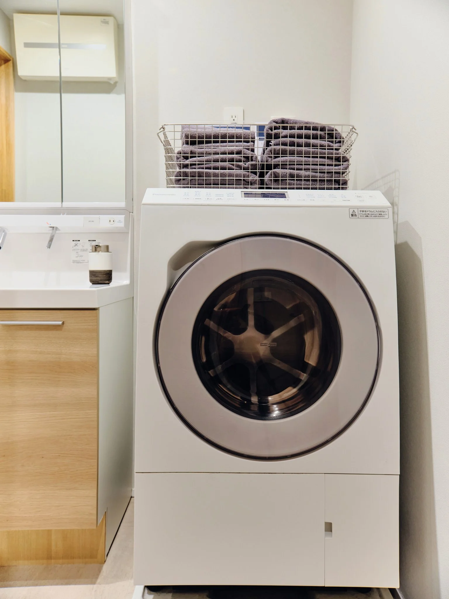 Washing machine with a wire basket on top filled with folded dark gray towels, located in a laundry room.