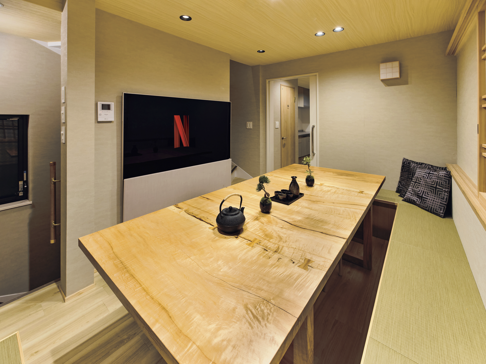 A cozy dining room with a large wooden table, a built-in bench with cushions, a TV with a Netflix logo, and decorative vases with plants.