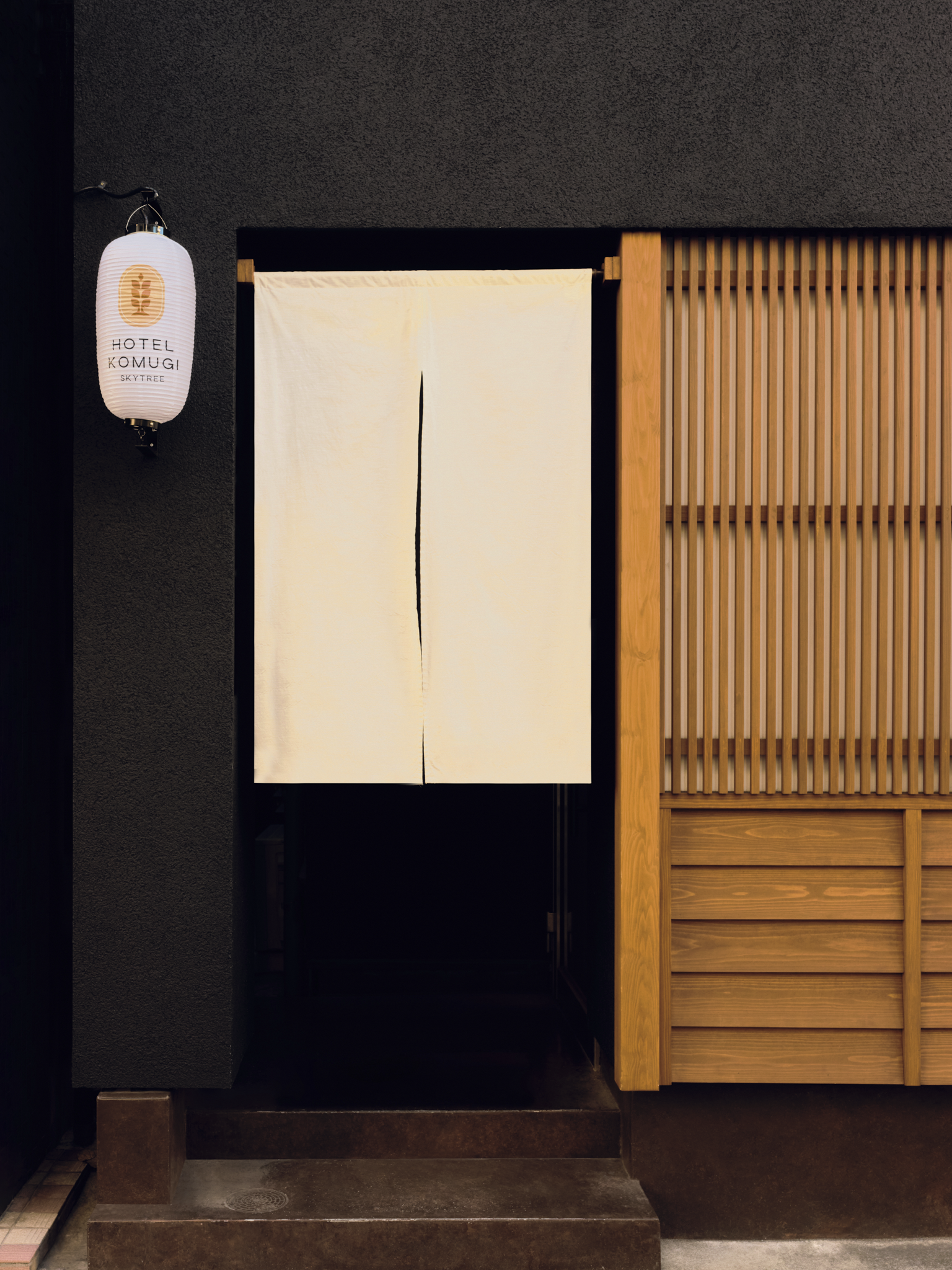 Entrance to a Japanese-style building with a white noren curtain, black background, wooden accents, and a paper lantern labeled 'Hotel Komugi Sky Tree'.