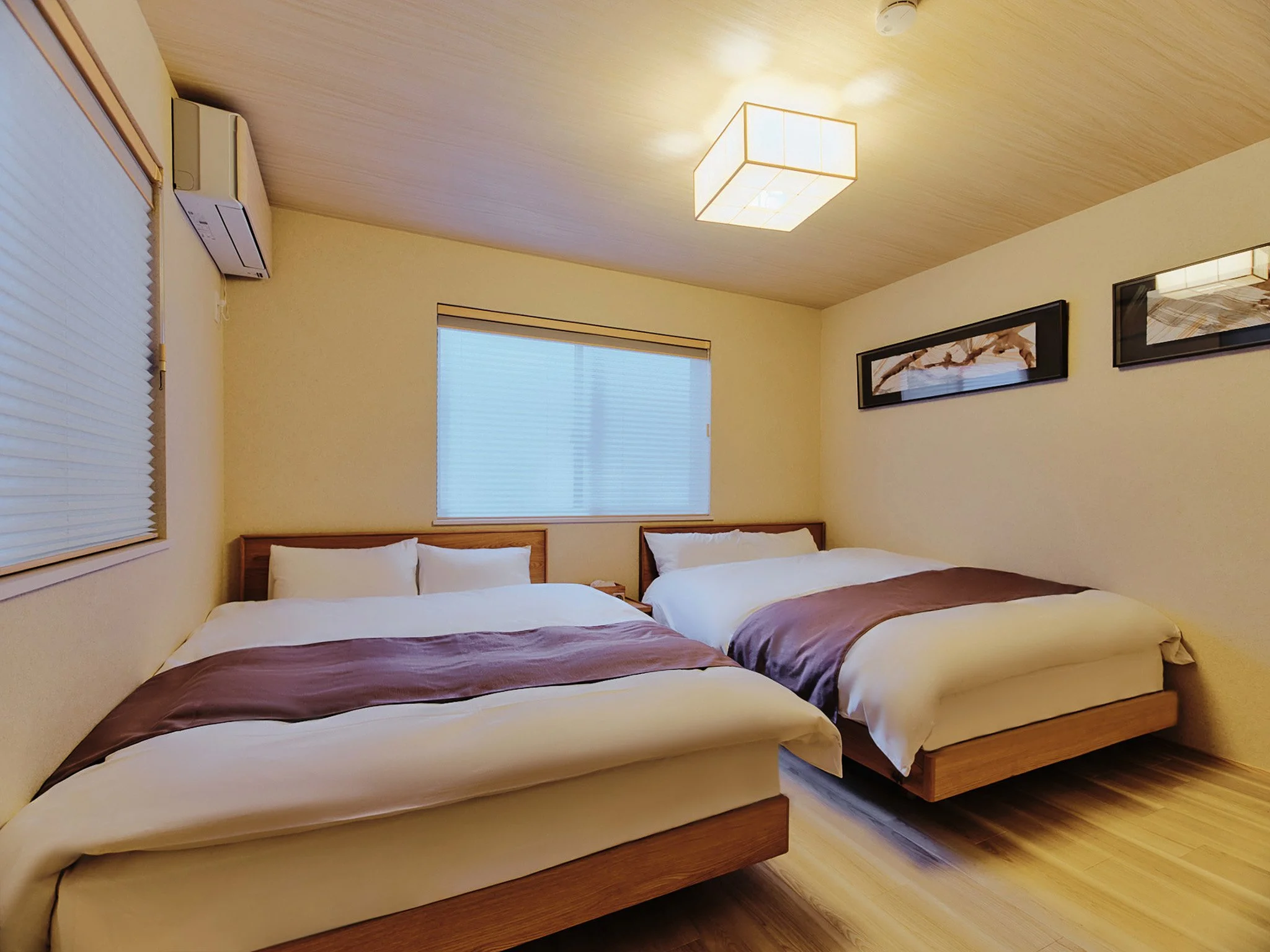 A cozy hotel room with two beds, wooden headboards, a window with closed blinds, two framed pictures on the wall, a ceiling light fixture, and a wall-mounted air conditioner.