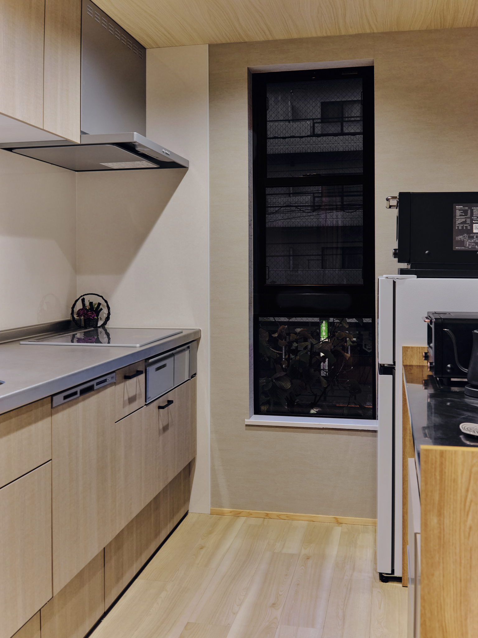 A modern kitchen with light wood cabinets, a stovetop, stainless steel range hood, small decorative basket, and a black-framed window showing an apartment balcony outside.