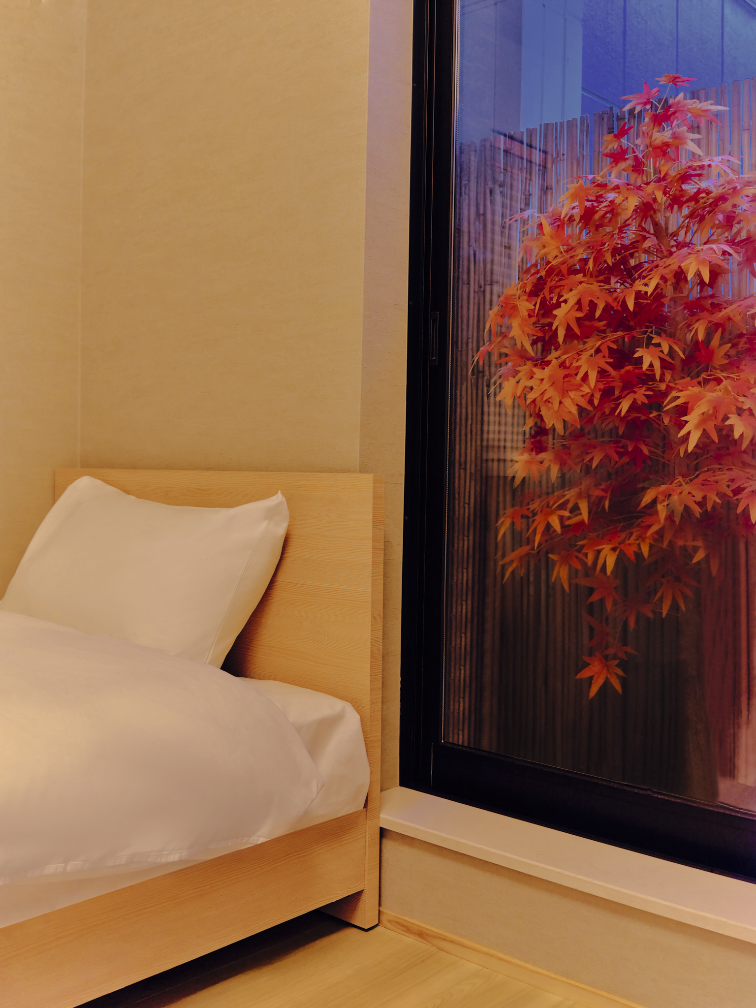 Interior of a bedroom featuring a wooden bed with white bedding and a pillow, and a large window showcasing an orange-red autumn tree outside.