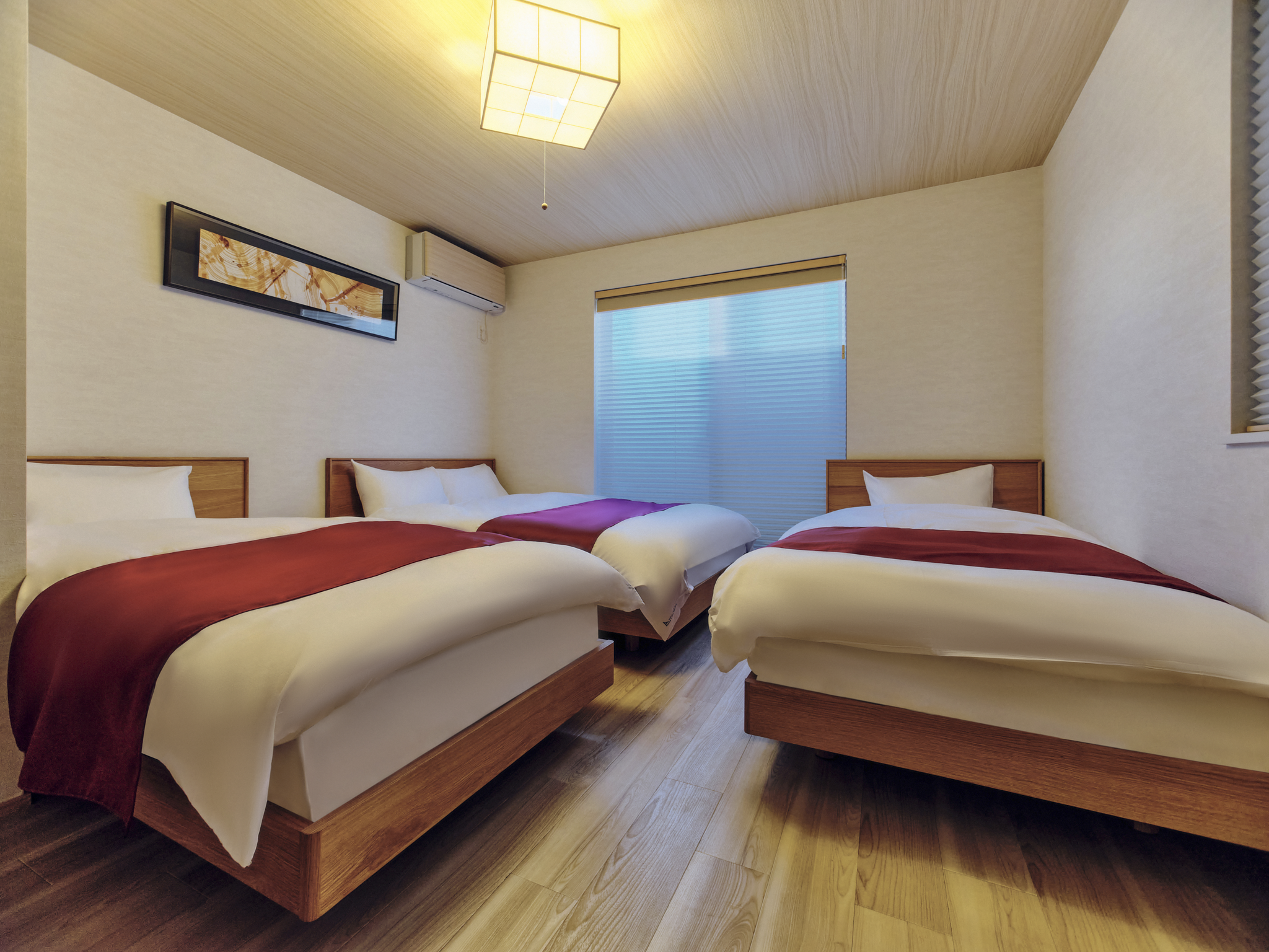 A hotel room with three beds, each with white bedding and red runners, a framed art piece hanging on the wall, a window with blinds, wooden flooring, and an air conditioning unit.