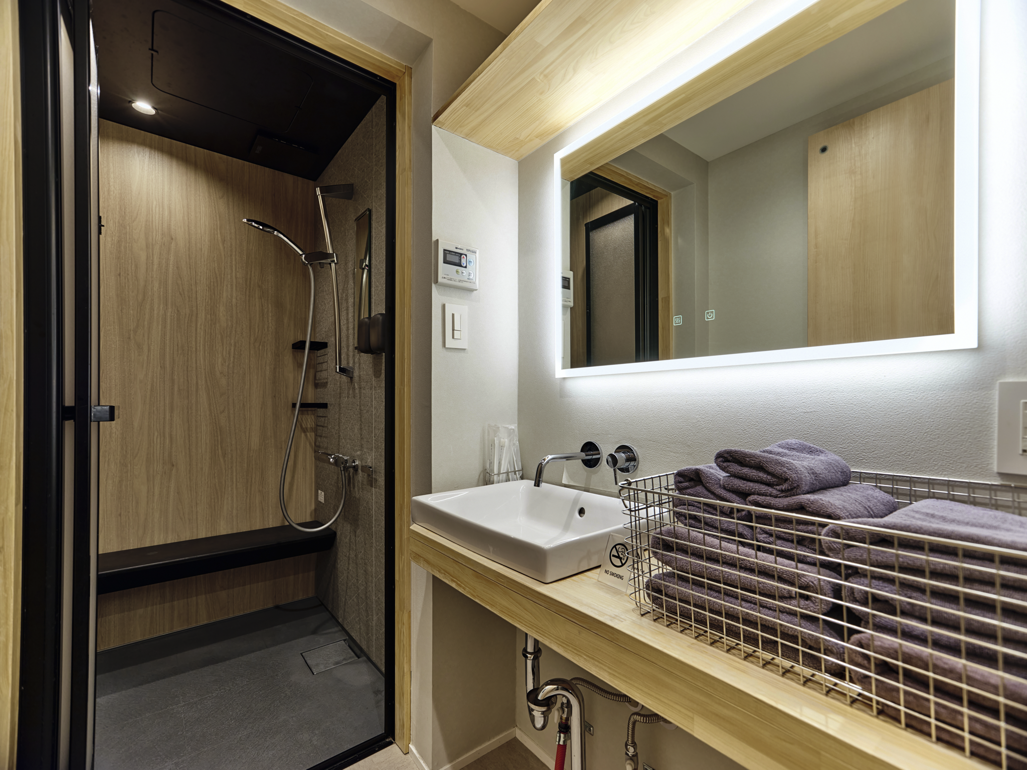 Modern bathroom with a wooden shower area, a large illuminated mirror, a white sink, and a wire basket filled with folded purple towels.