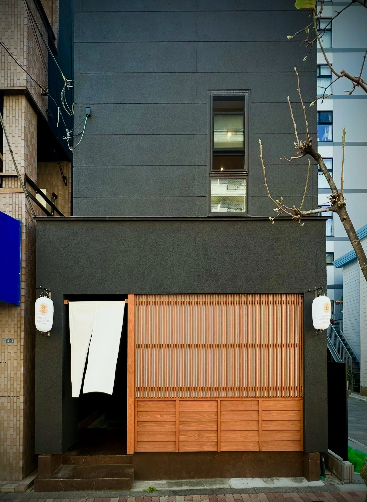 A modern building façade with a dark gray exterior, small vertical window, and a traditional Japanese entrance with a white noren curtain, wooden sliding door, and two white paper lanterns.