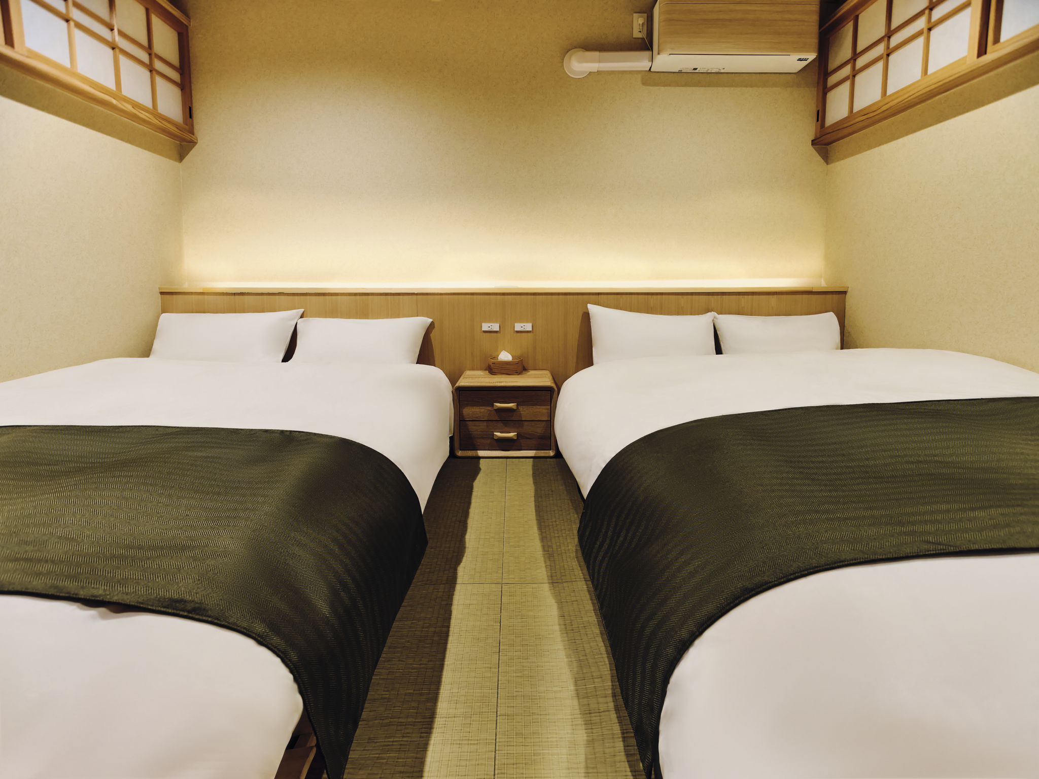 A hotel room featuring two large beds with white linens and dark green bed runners, a wooden nightstand with a tissue box, beige walls, and a ceiling-mounted air conditioning unit.
