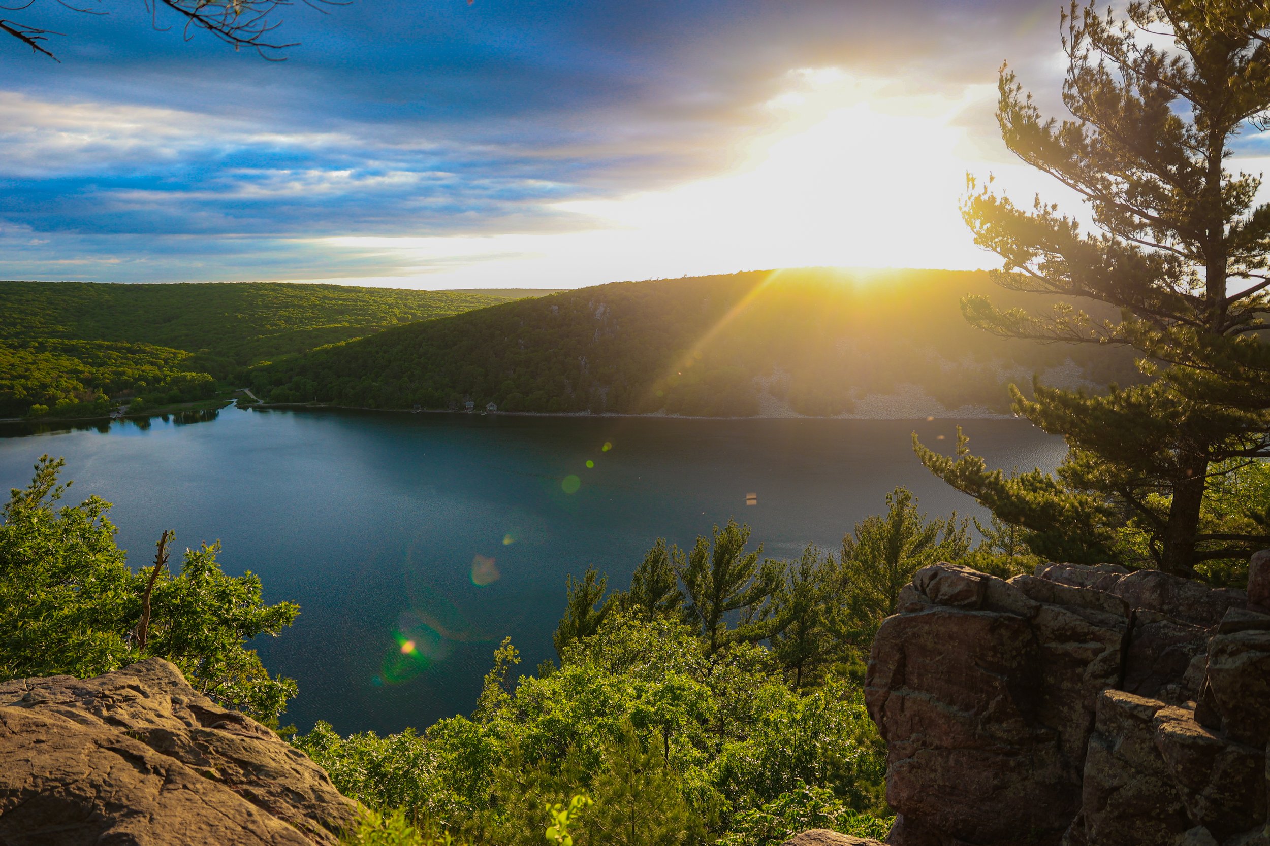 Sunset over a lake surrounded by green hills and trees, with rocks and foliage in the foreground.