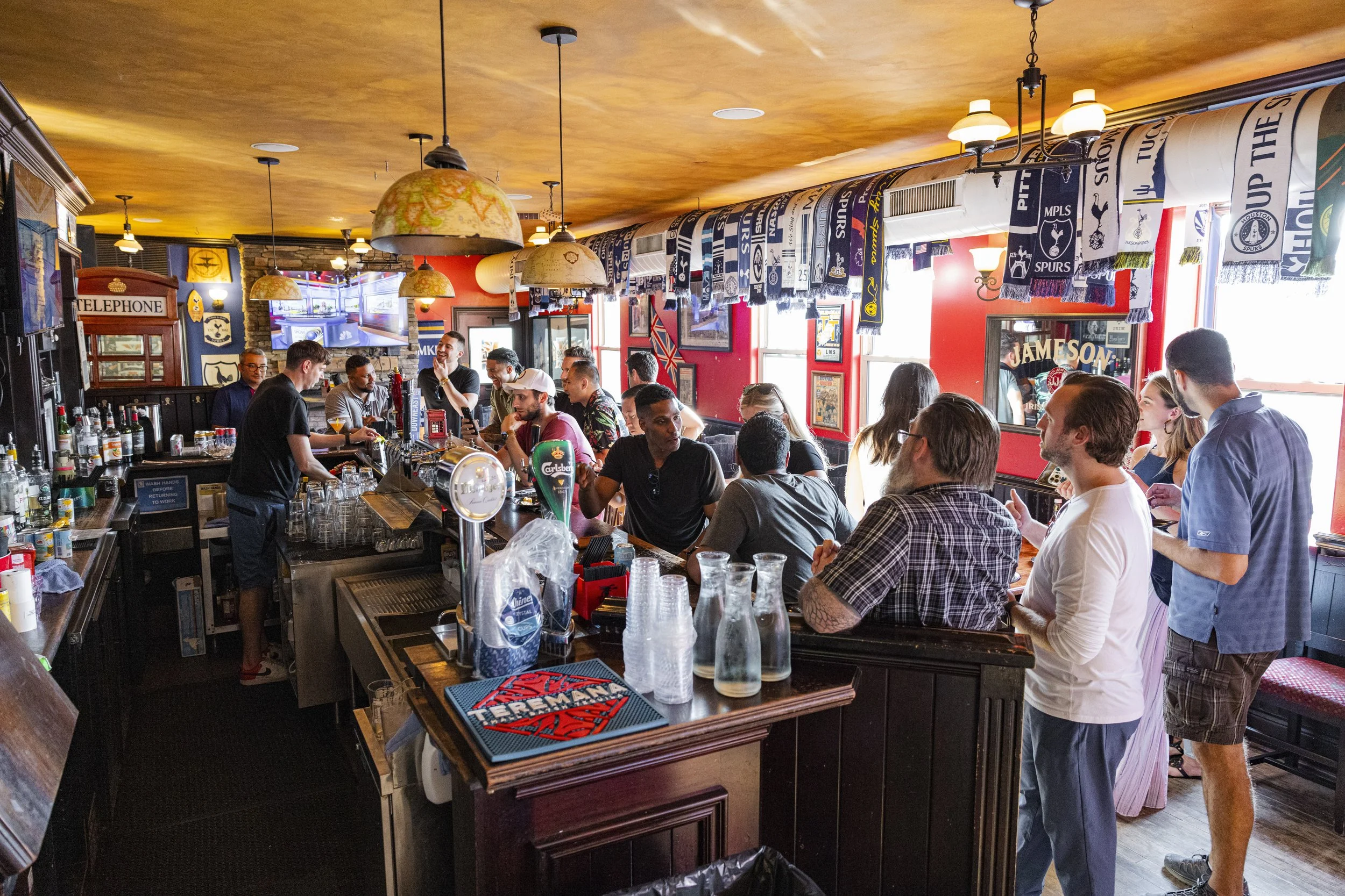 Inside a lively bar with people sitting at the counter and standing, talking, with sports scarves hanging above and red-painted walls decorated with framed pictures and signs, and a bartender preparing drinks.