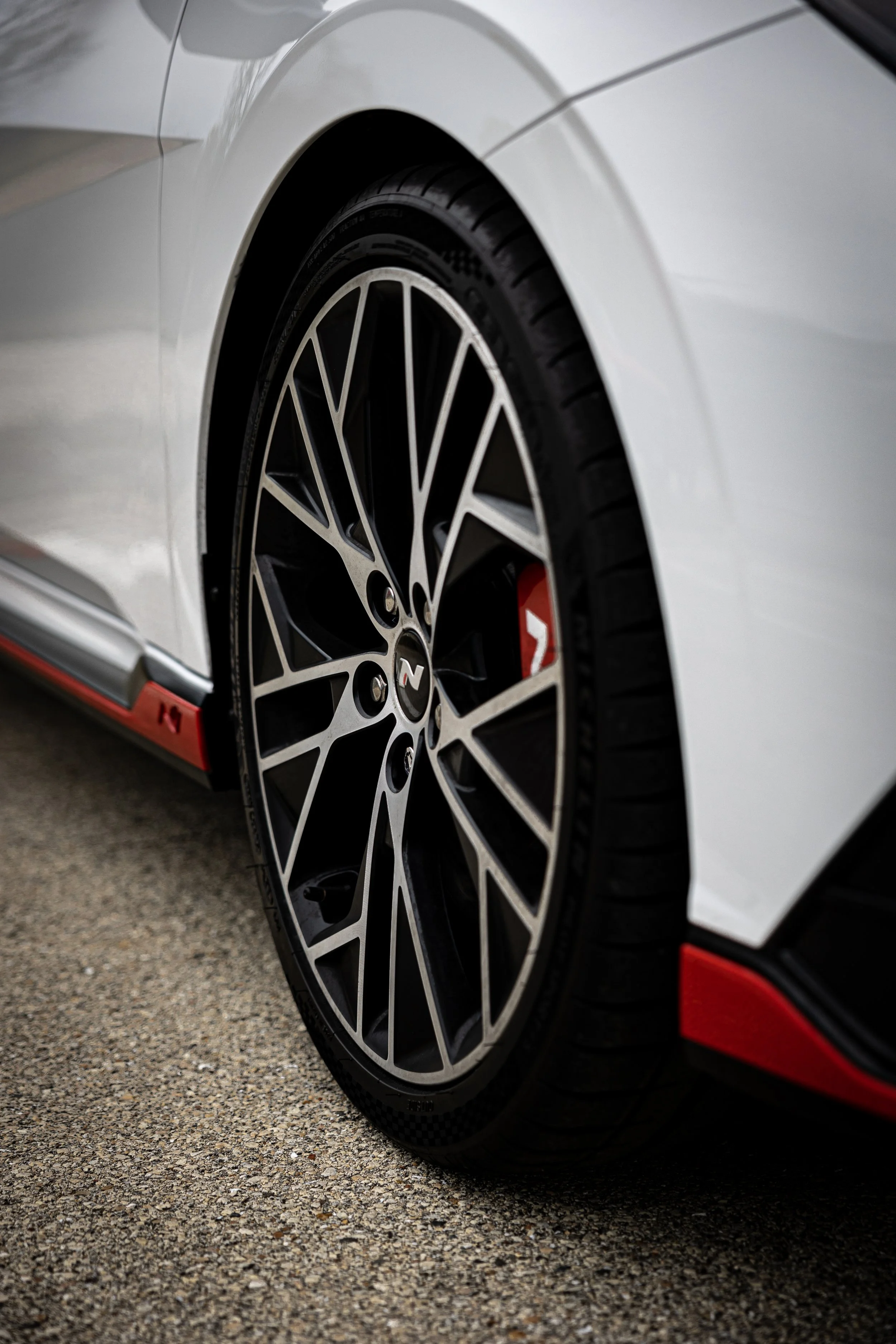 Close-up of a white sports car wheel with black and silver rim design and a red brake caliper, parked on a textured asphalt surface.