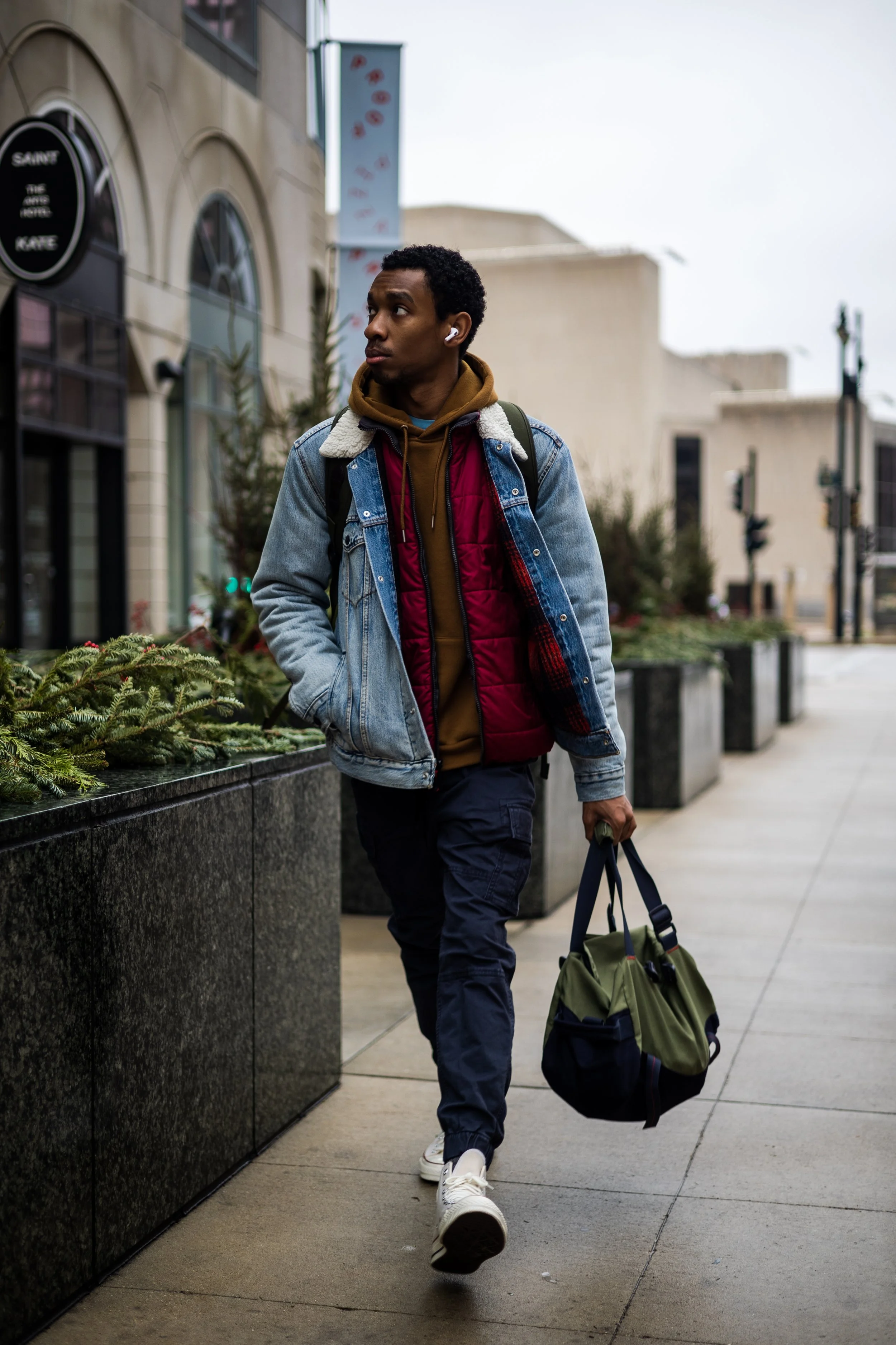 A young man walking on a city sidewalk, carrying a duffel bag, wearing headphones, a layered jacket, and casual pants, with storefronts and streetlights in the background on an overcast day.