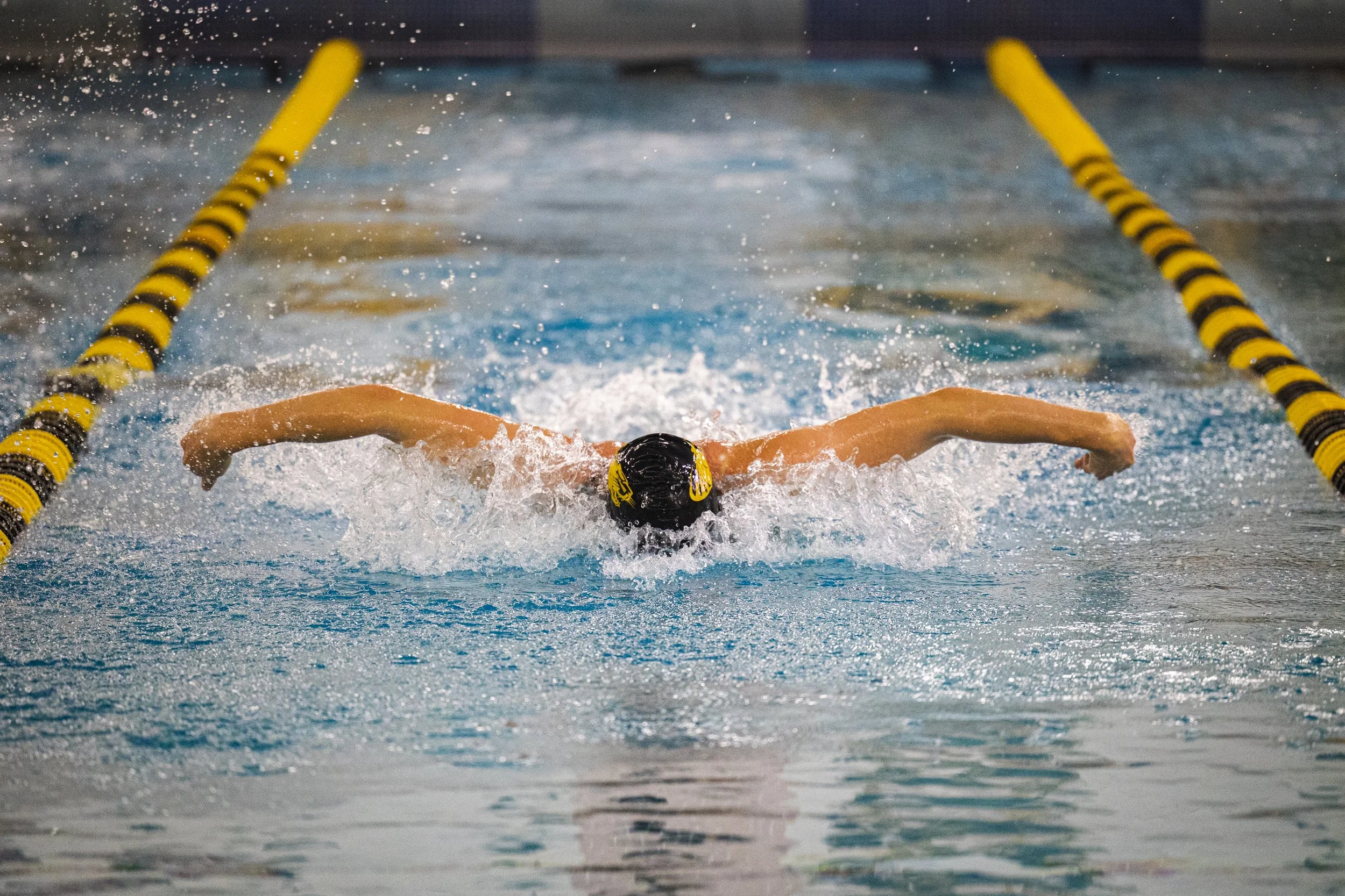 Swimmer in a pool performing the butterfly stroke, wearing a black swim cap with yellow accents, between yellow and black lane dividers.
