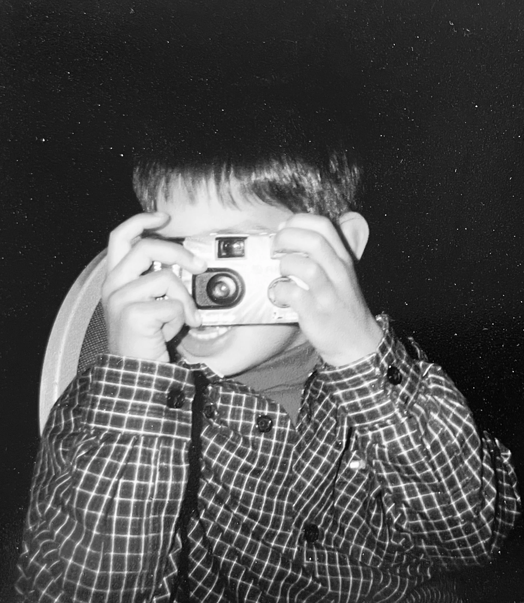 Child taking a photo with a camera, wearing a checkered shirt, black background.