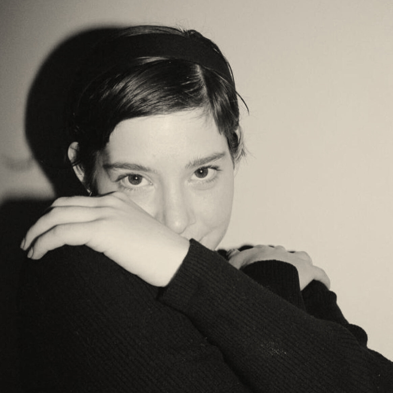 Black and white portrait of a young woman with short dark hair, resting her face on her crossed arms, looking directly at the camera.