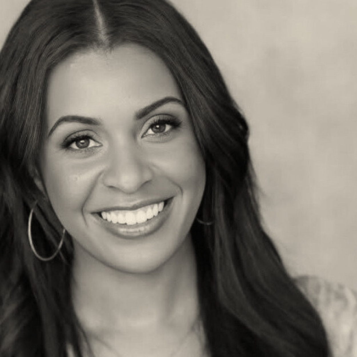 A woman with long dark hair, smiling, wearing hoop earrings, in a black and white portrait.