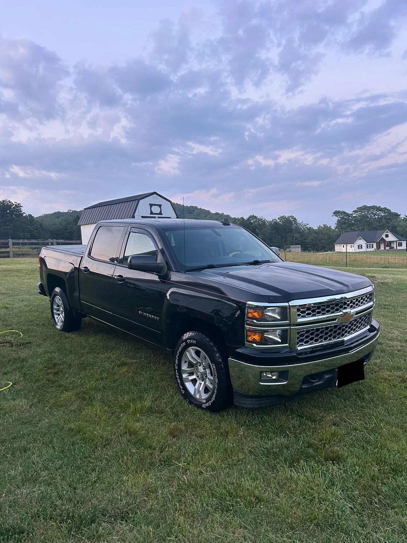Black Chevrolet Silverado pickup truck parked on grass with a small white shed with a black roof and a house in the background under a cloudy sky.