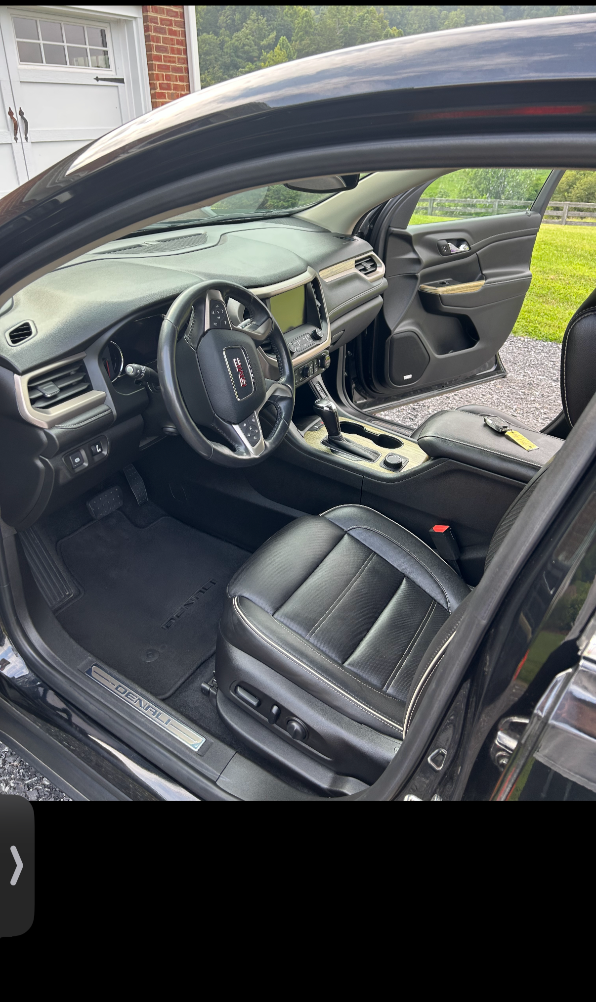 The interior of a black GMC vehicle showing the driver's seat, steering wheel, dashboard, center console, and open front passenger door, with a green outdoor landscape in the background.