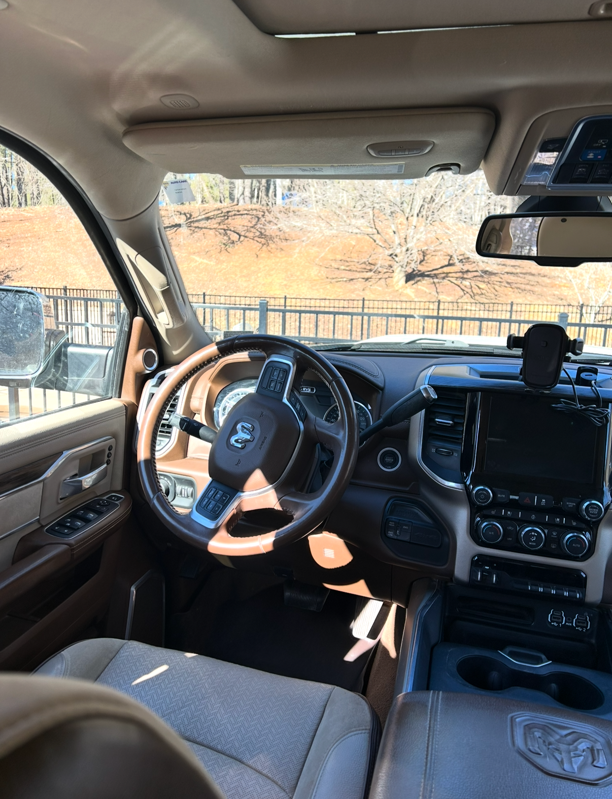 Interior of a beige and brown Ram truck with a steering wheel, dashboard, and touchscreen display, parked outdoors near a fence and trees.