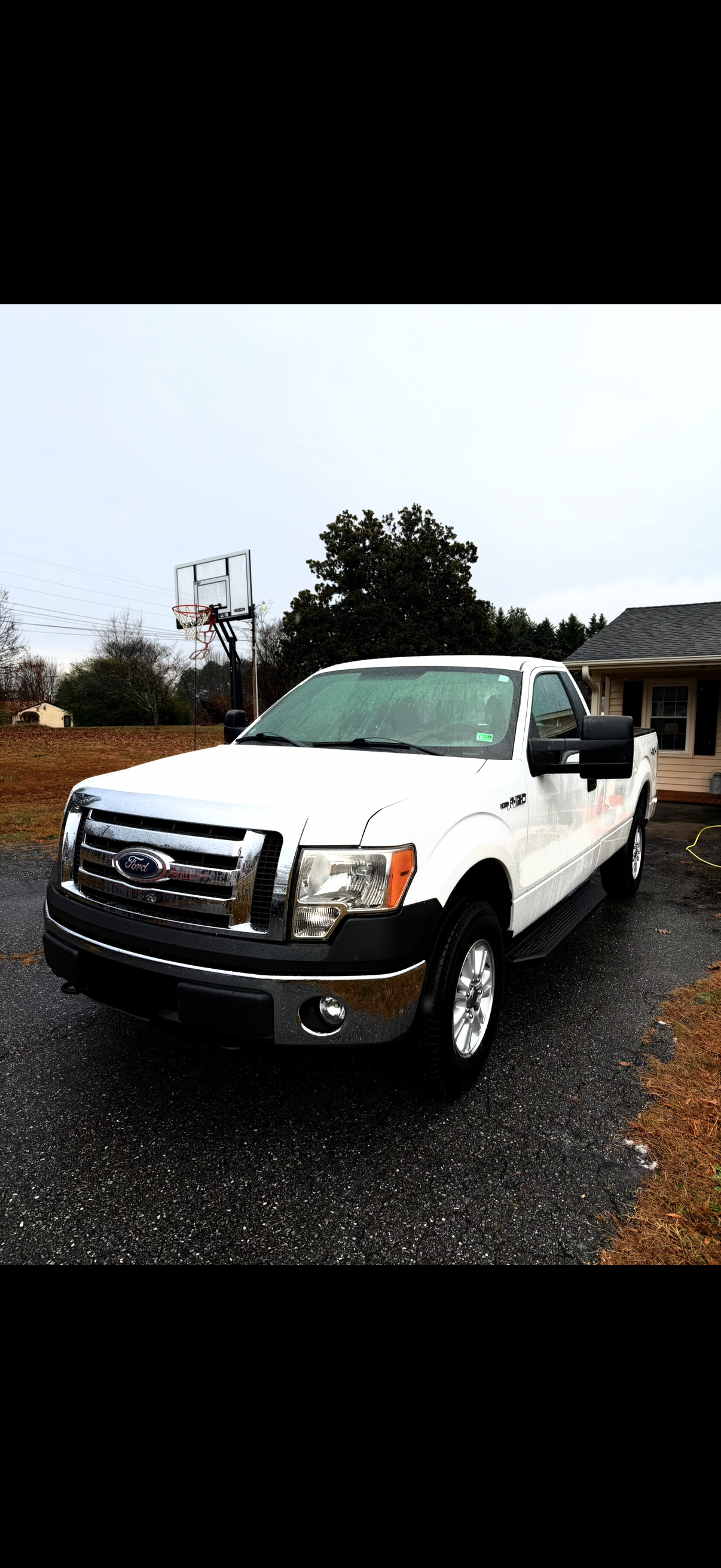 White Ford pickup truck parked on wet driveway with a basketball hoop in the background and a house to the right. Rain is visible on the truck's windshield.