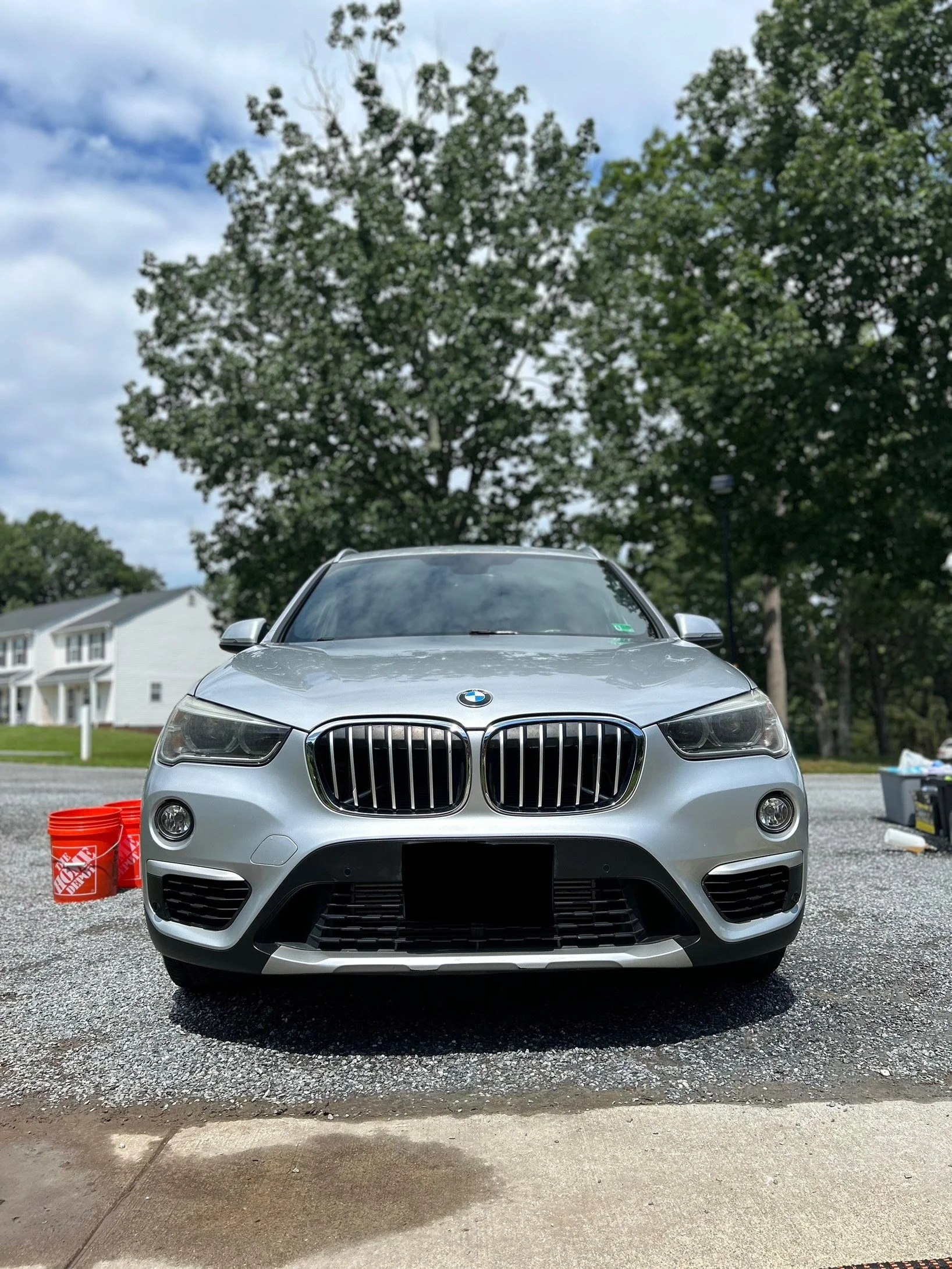 Front view of a silver BMW SUV parked outdoors on a gravel driveway, with trees, blue sky, and white houses in the background.