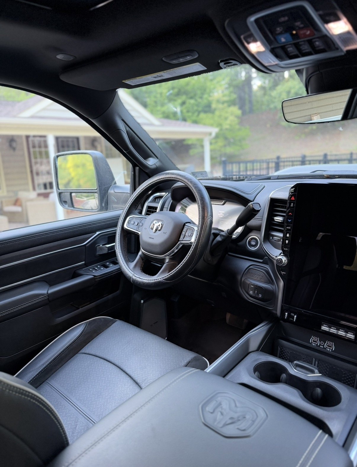 Interior of a pickup truck showing the steering wheel, dashboard, and front passenger seat; outside view of a house and greenery through the windows.