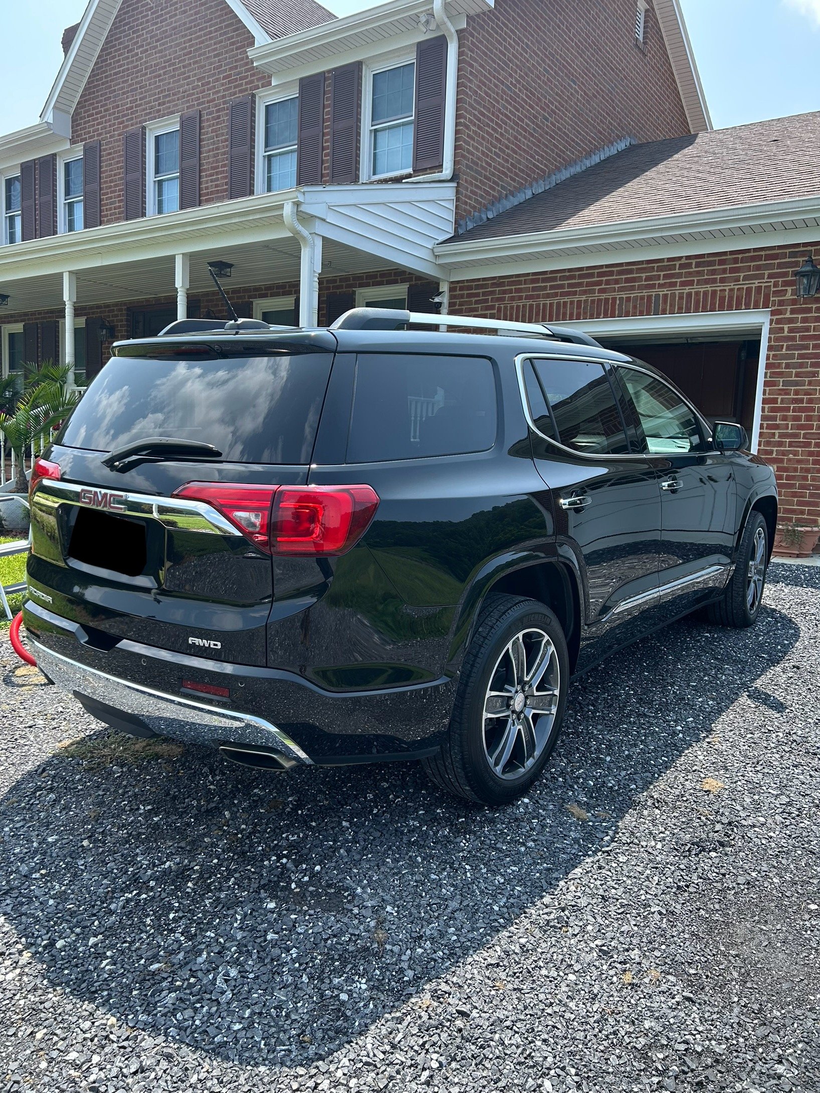 A black GMC SUV parked on gravel driveway in front of a brick house with white trim and brown shutters.