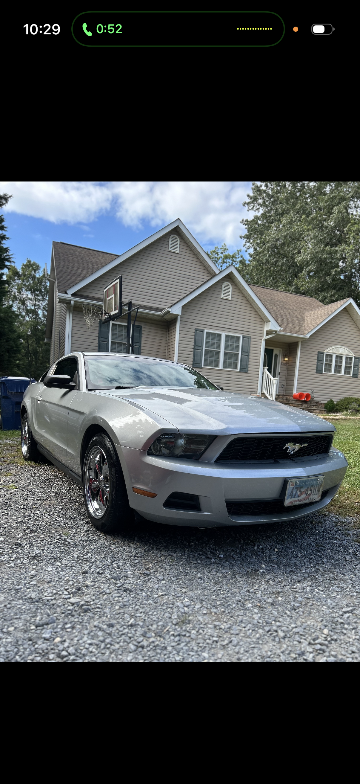 A silver Ford Mustang parked on a gravel driveway in front of a beige house with gray shutters. The house has a small porch with white railings, a front door, and multiple windows. There is a basketball hoop attached to the house and a large tree with green leaves in the background.
