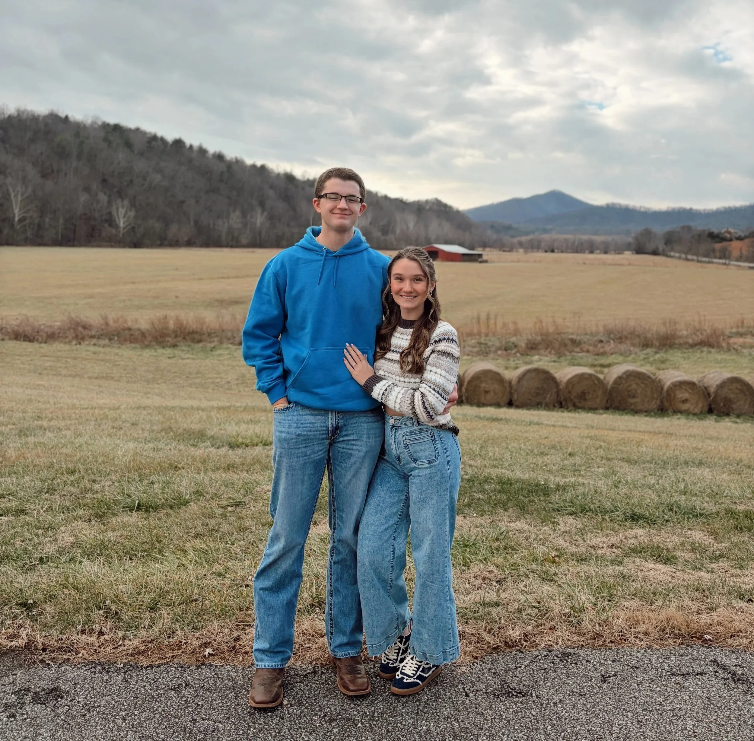 A young man and woman standing together outdoors in a rural field with a barn and hills in the background. The man is wearing a bright blue hoodie and jeans, and the woman is wearing a patterned sweater and jeans. They are smiling at the camera.