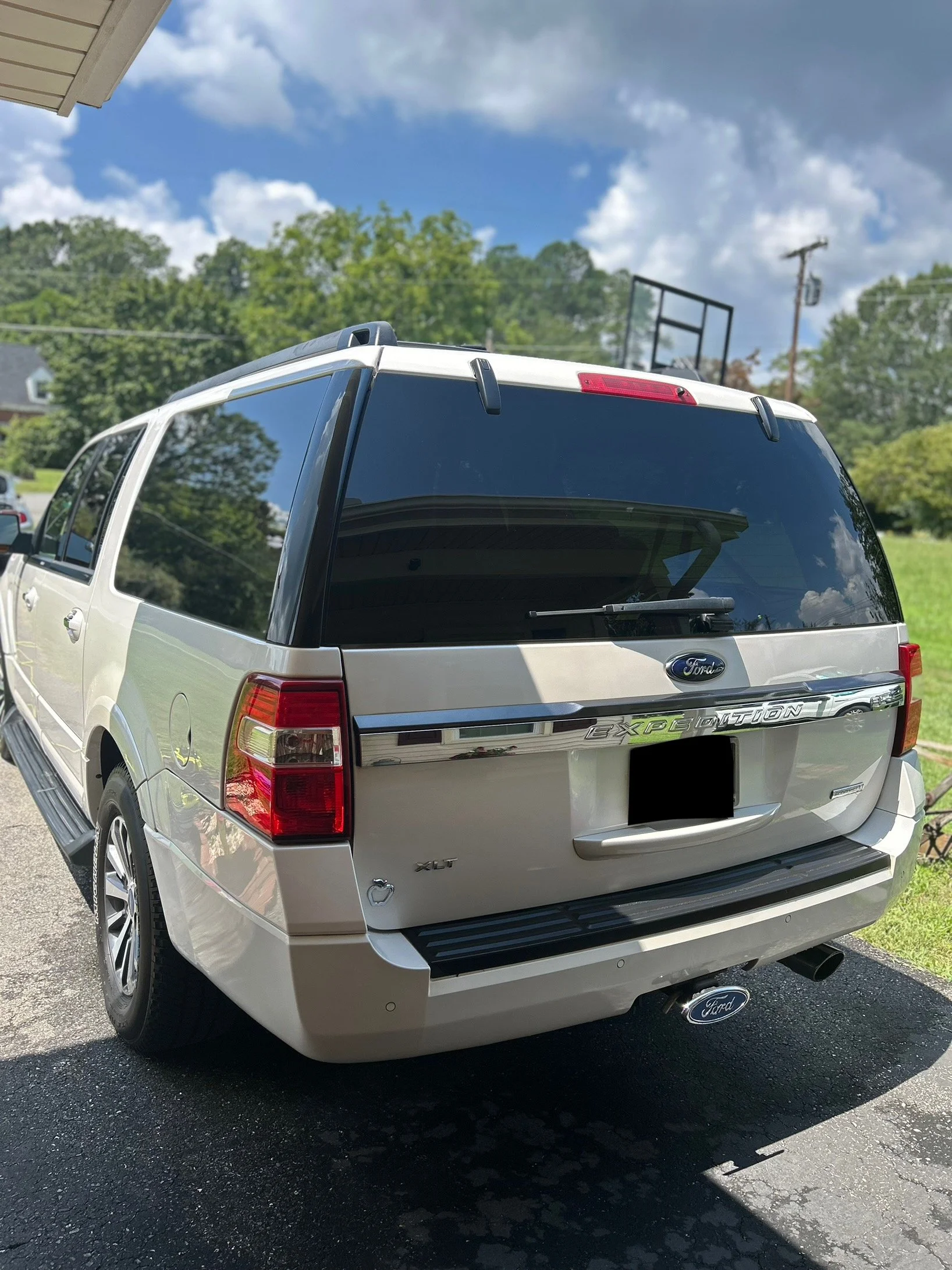 Rear view of a silver Ford Expedition SUV parked on a driveway on a sunny day with trees and a grassy area in the background.