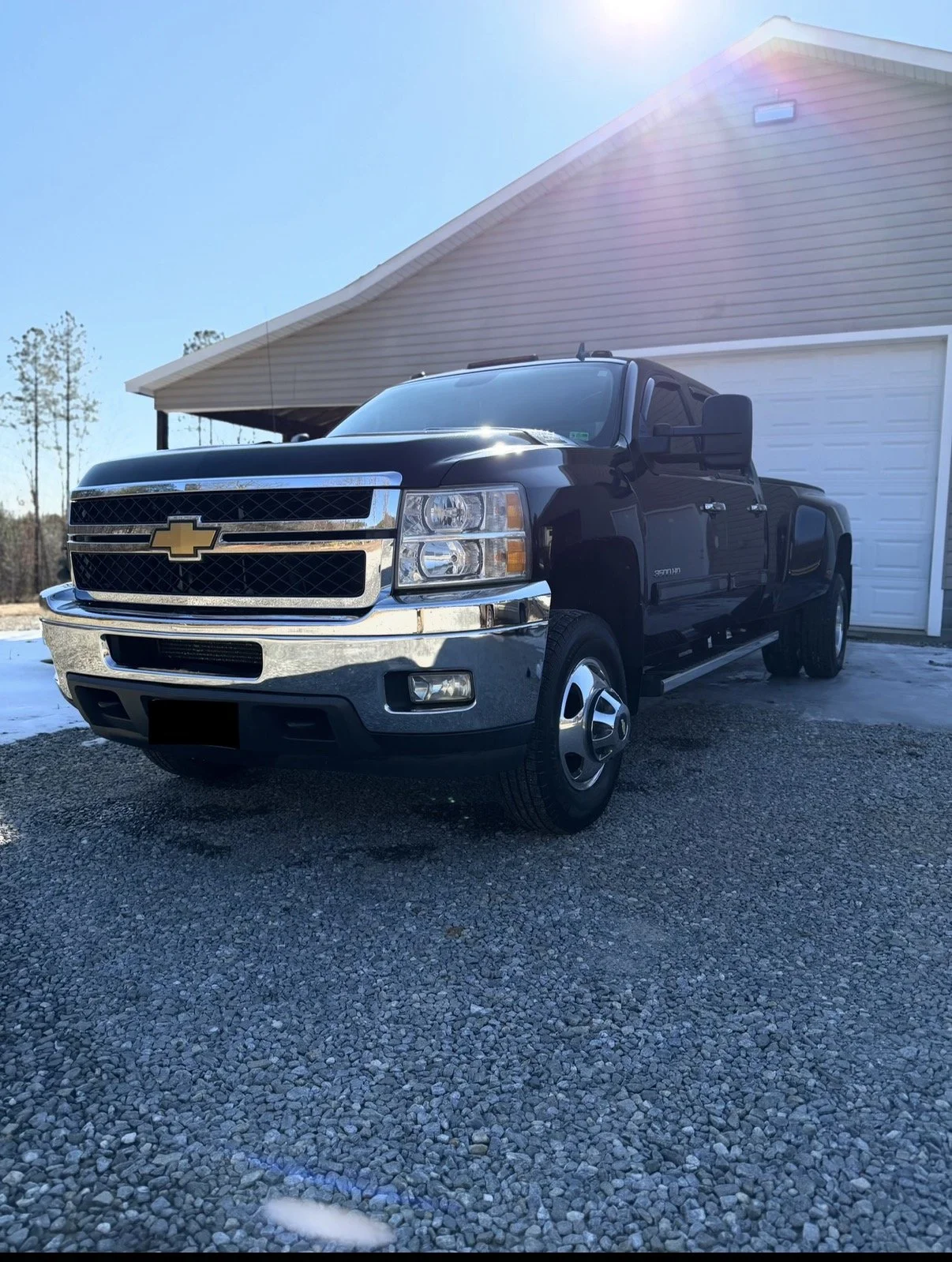 Black Chevrolet Silverado pickup truck parked outside a house with a white garage door on a gravel driveway, under a bright blue sky.