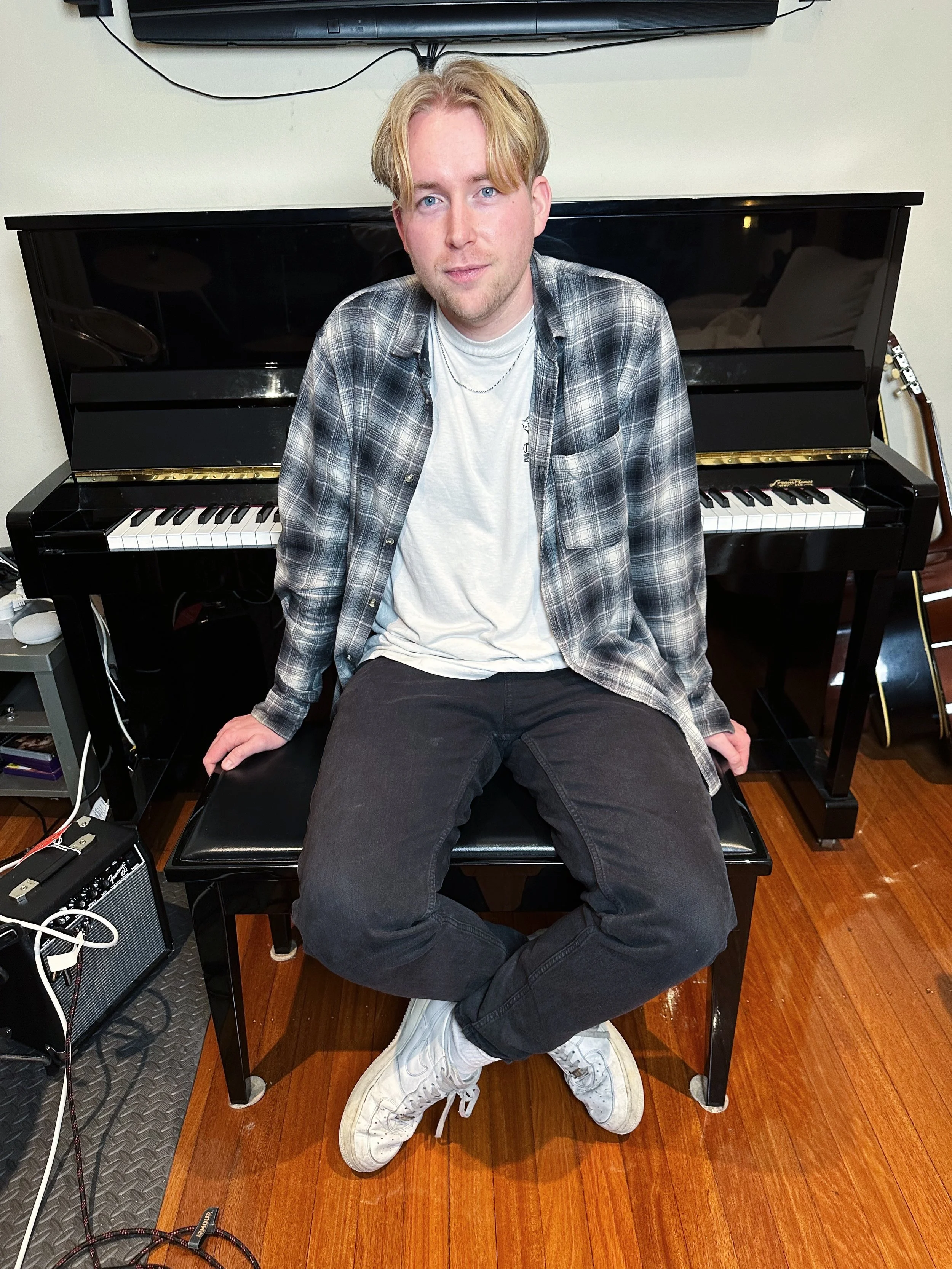 A young man with blonde hair sitting on a piano bench in front of a black upright piano in a room with wooden flooring. He is wearing a white t-shirt, a plaid shirt, black jeans, and white sneakers.