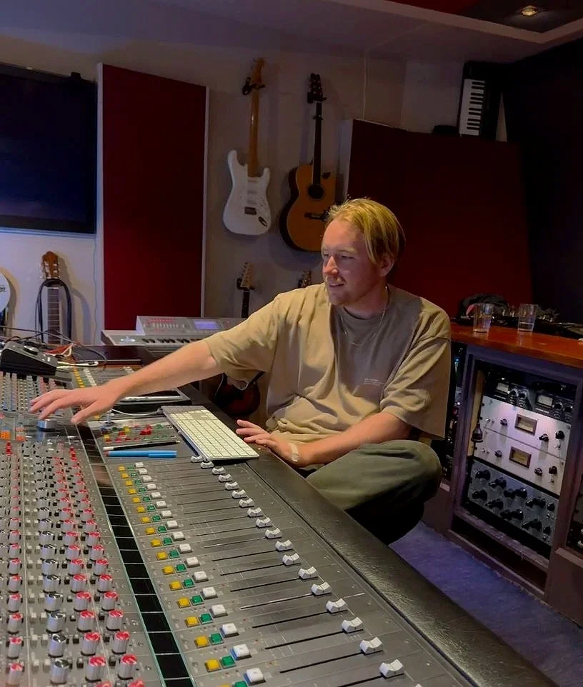 A person working at a music recording studio mixing console with guitars hanging on the wall behind him.