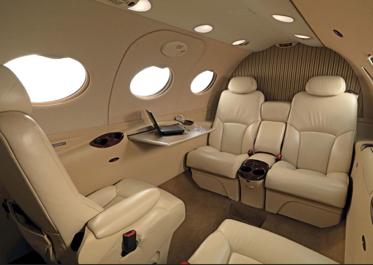 Interior of a private jet with beige leather seats, three seats on one side, two on the opposite, a small table, windows, and a laptop.