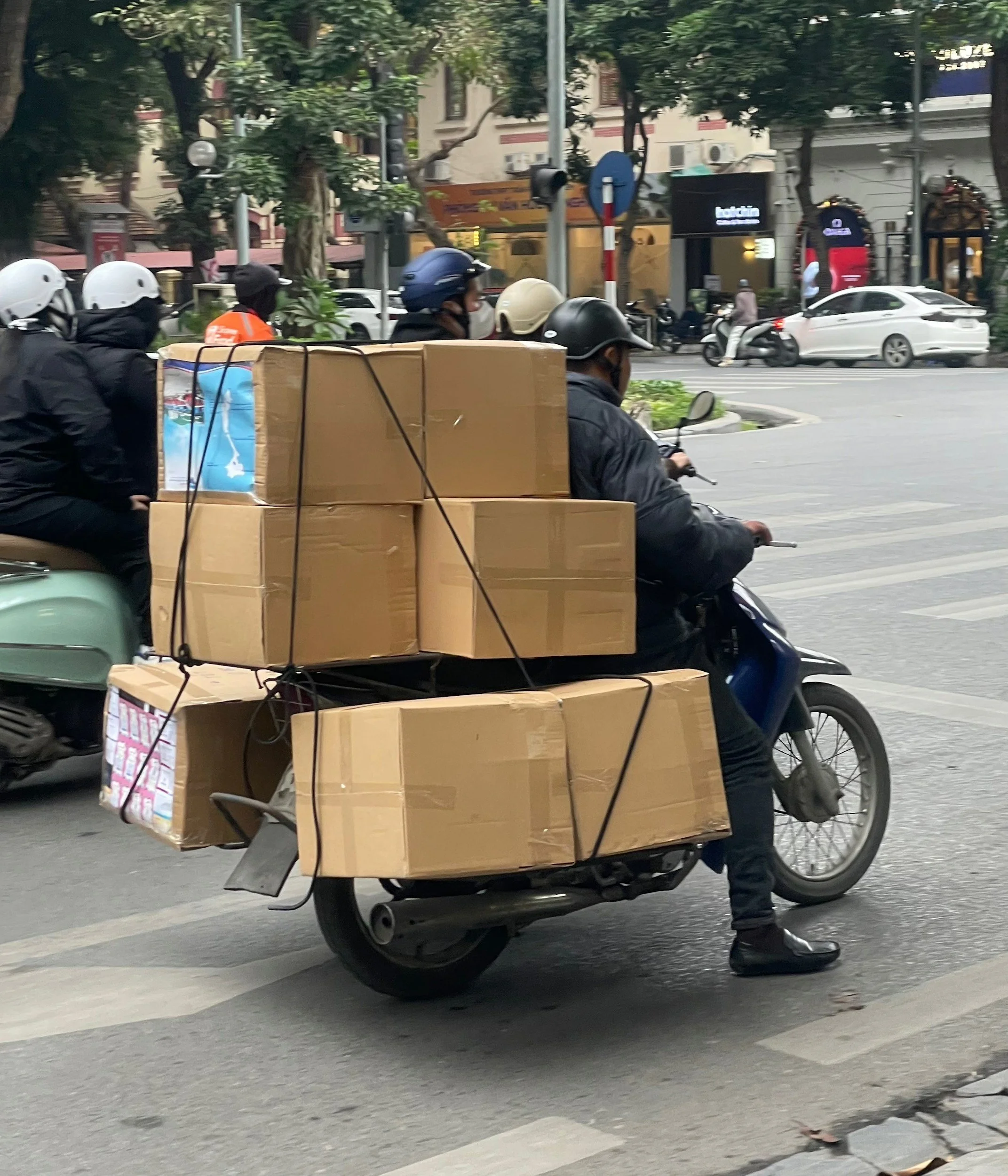 A person riding a motorcycle on an urban street carrying numerous cardboard boxes strapped to the back of the motorcycle.