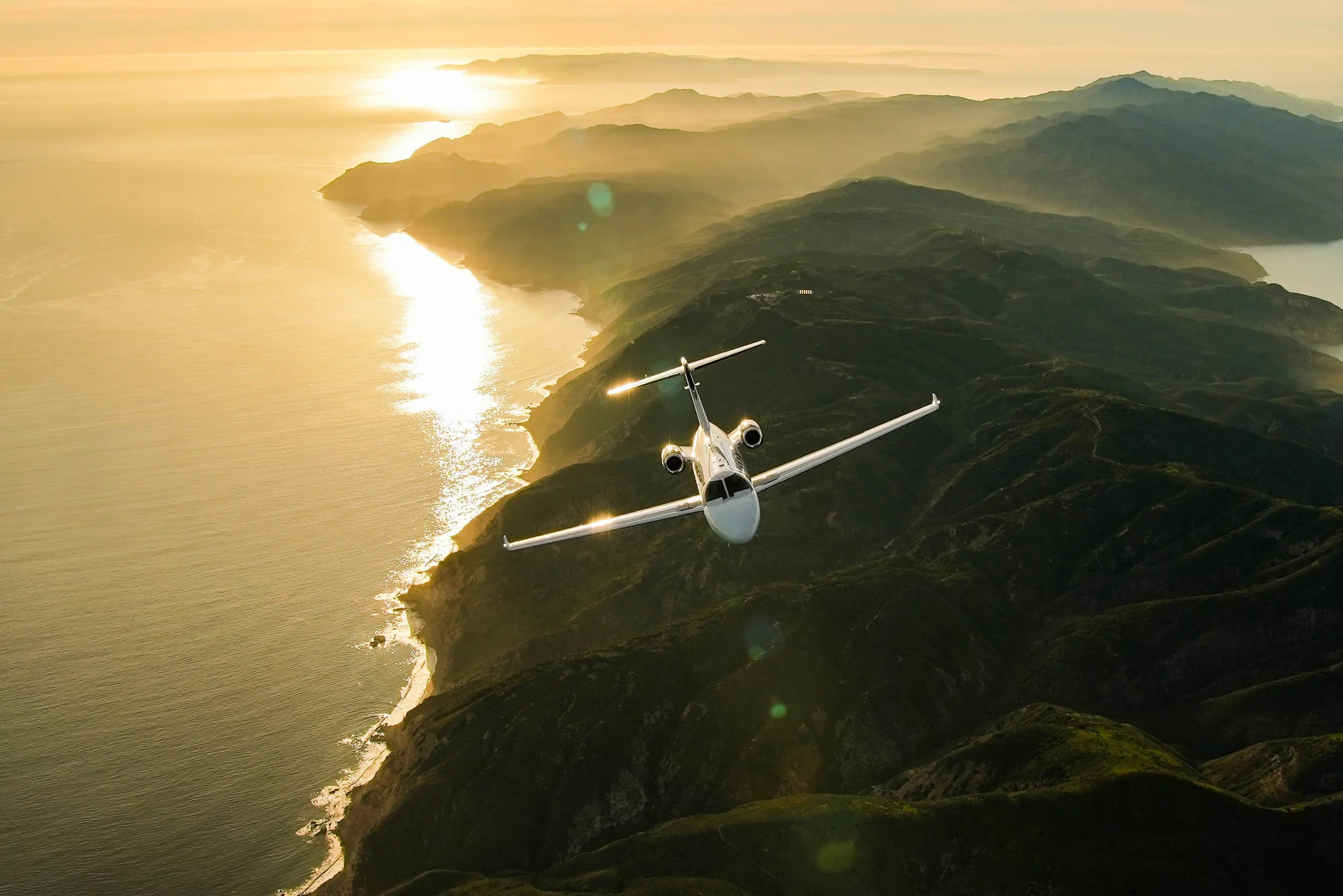 A small jet flying over a mountainous coastline during sunset with the ocean on the left and green hills on the right.