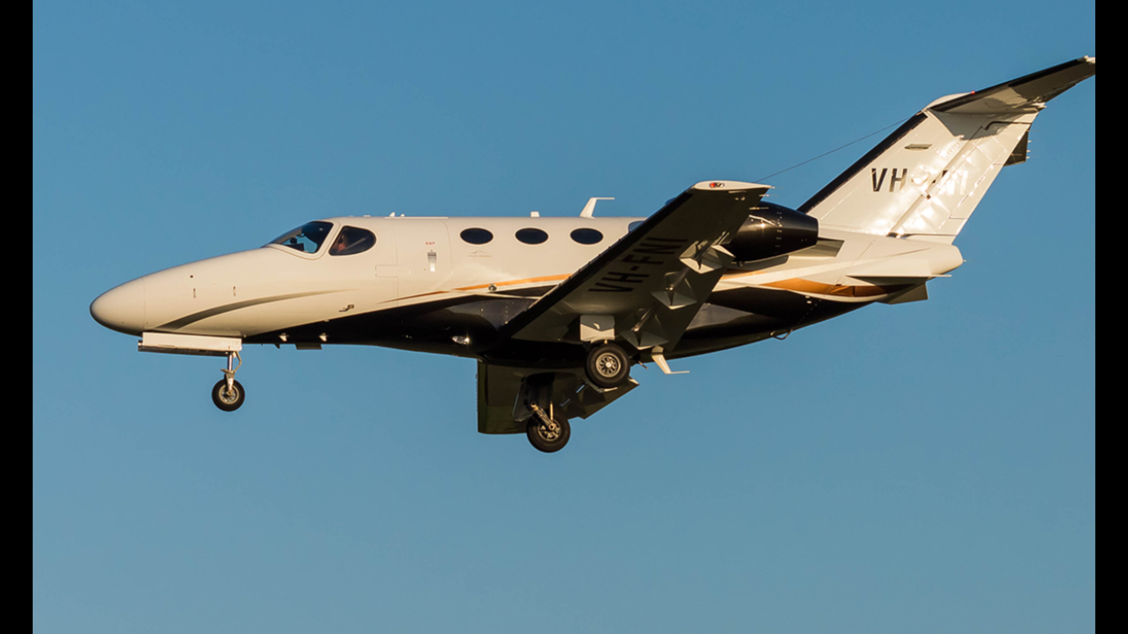 A white private jet flying against a clear blue sky with its landing gear extended.