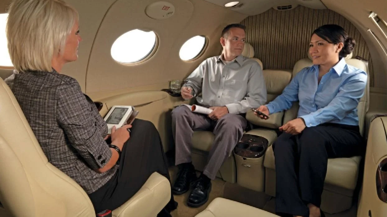 Three business professionals seated inside a private jet, engaging in conversation, with two of them holding devices and the third holding a script or document.