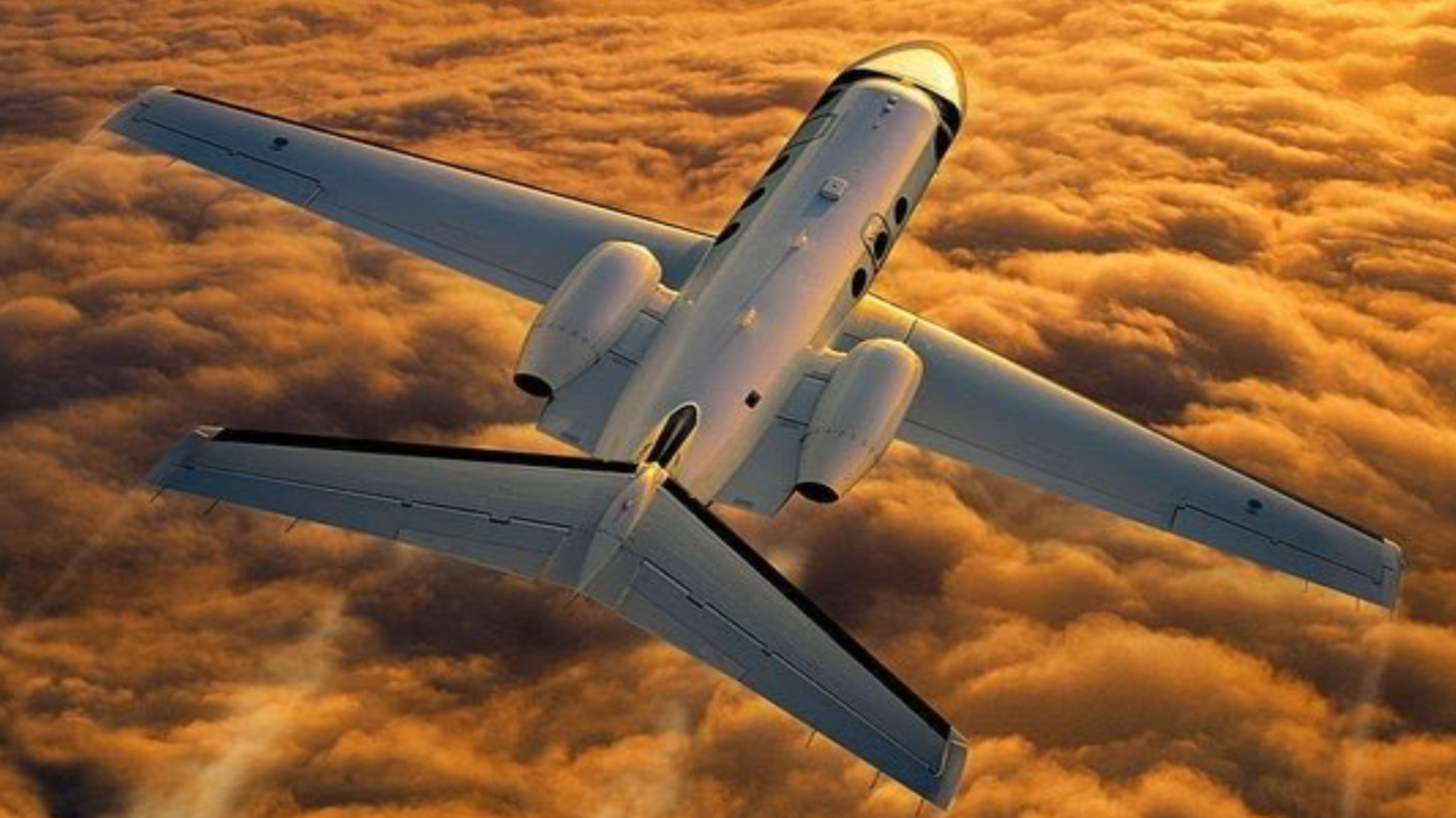 An aerial view of a white jet airplane flying above orange-tinted clouds during sunset or sunrise.