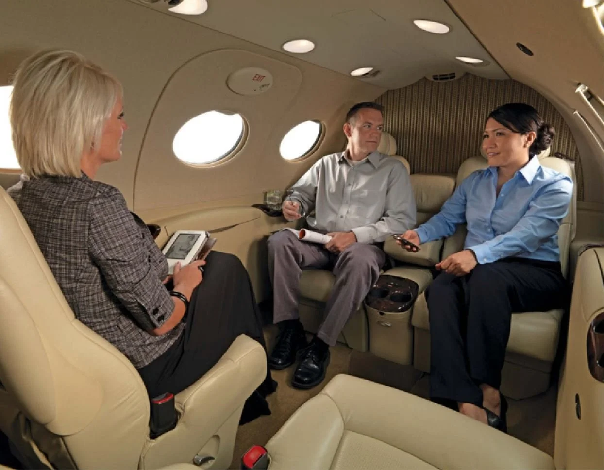 Three business professionals seated inside a private jet, engaged in conversation. The woman on the left is holding a notebook, the man in the middle has a rolled-up document, and the woman on the right is holding a phone.