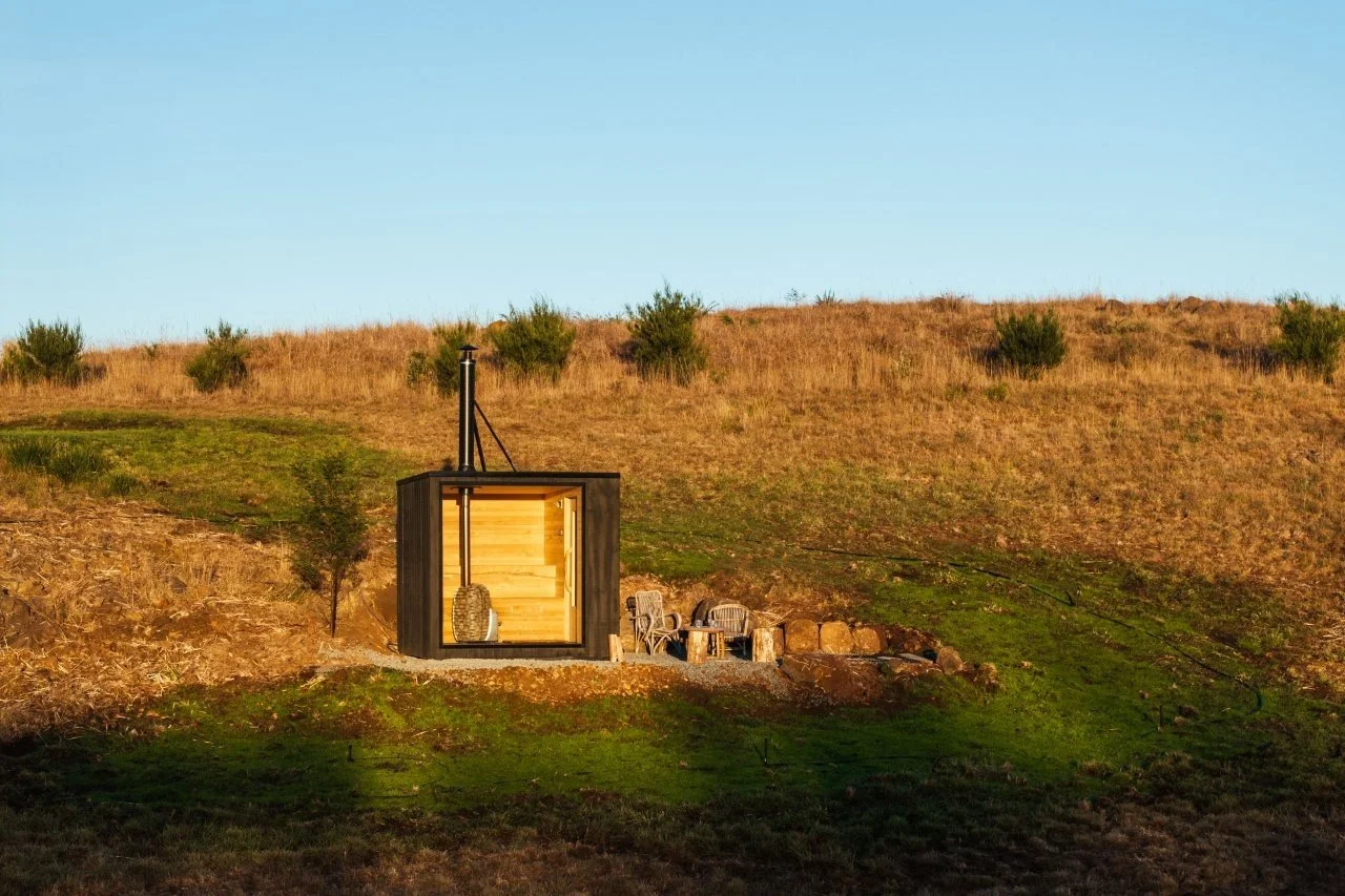A small modern cabin with a black exterior and large front windows, set on a grassy hillside with sparse trees and a clear blue sky in the background.