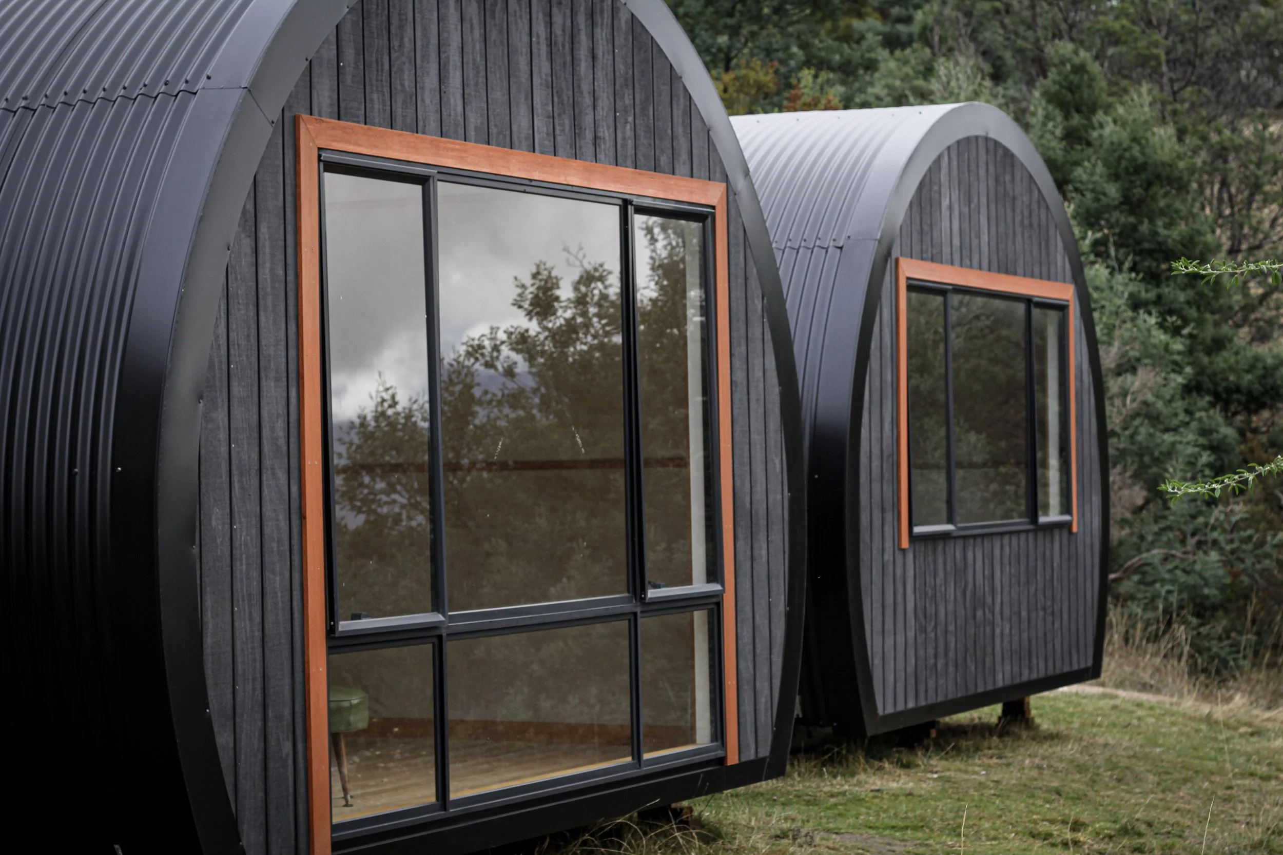 Two modern tiny houses with curved black exteriors and large sliding glass windows, situated outdoors on grassy terrain with trees in the background.