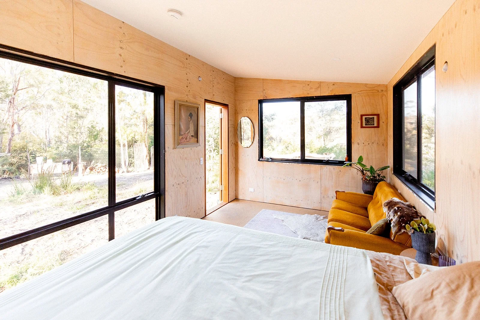 Room with large windows, a bed in the foreground, a yellow velvet sofa with a blanket, framed artwork on the walls, a mirror, plants, and a view of trees outside.