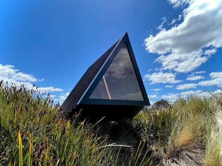 Modern black triangular glass building on grassy landscape against blue sky with clouds.