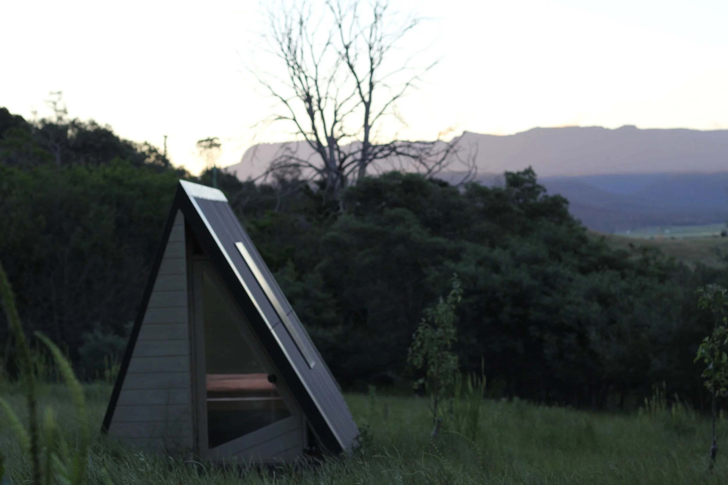 A modern, triangular A-frame tiny house with solar panels on the roof, situated on a grassy field with trees and mountains in the background at sunset.