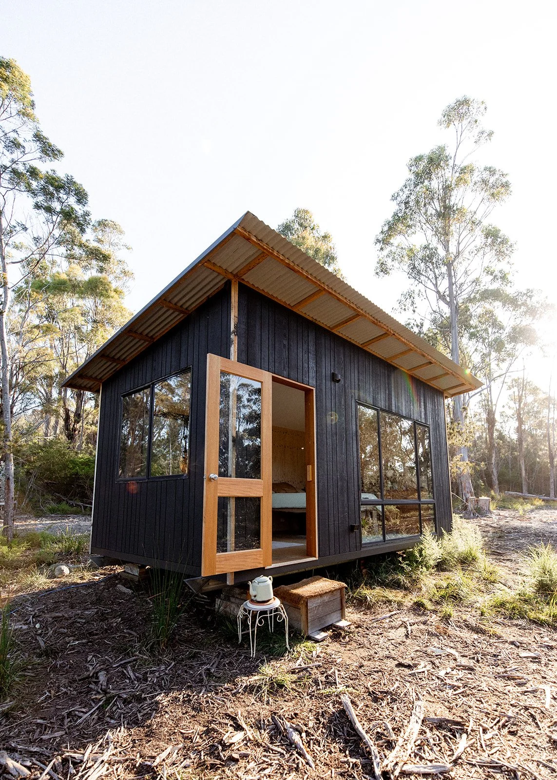 Small black cabin with large glass windows, wooden door, and raised foundation in a wooded area, with sunlight and trees in the background.