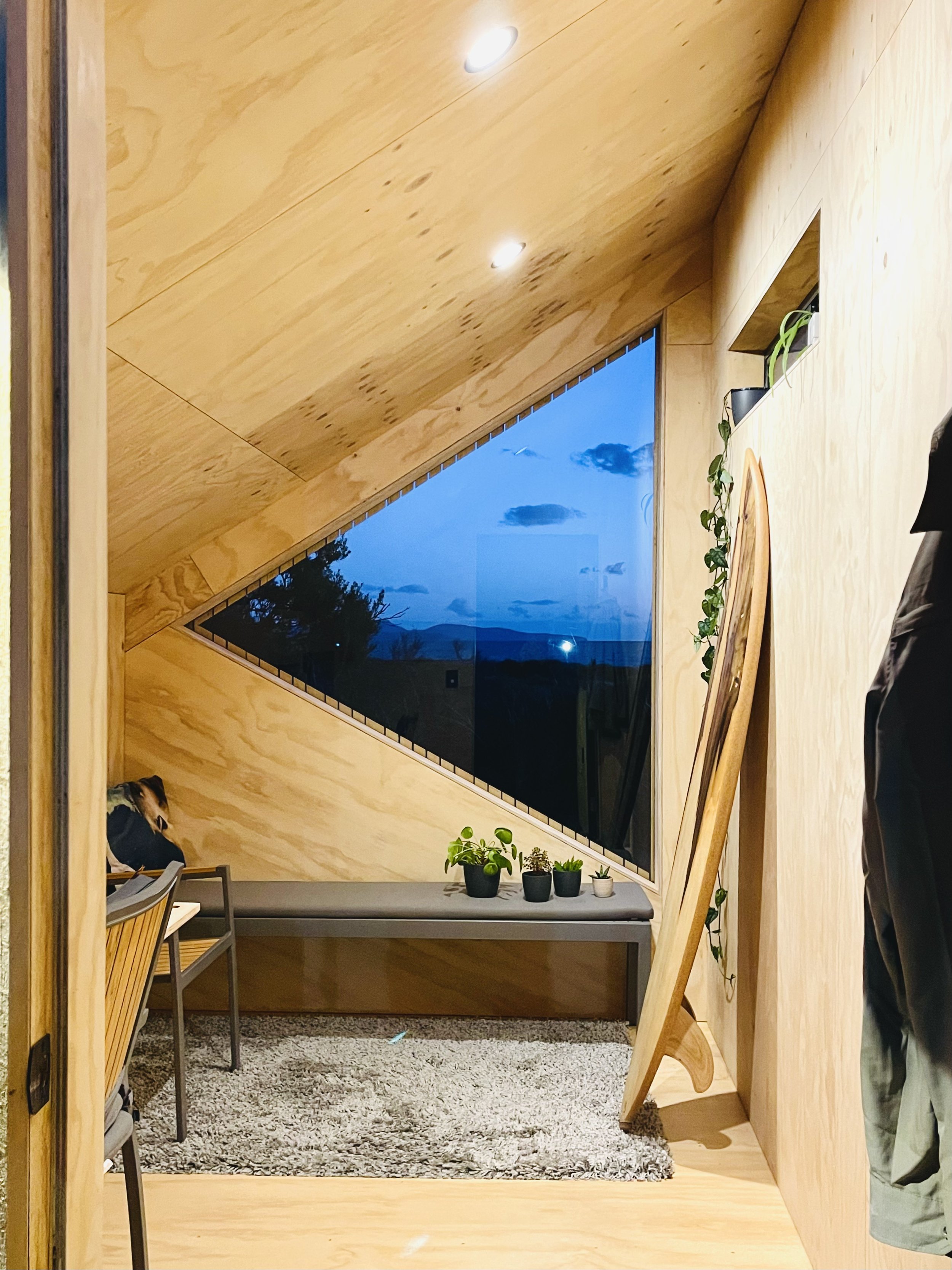 View of a cozy room with wood-paneled walls and ceiling, featuring a large triangular window showing a blue evening sky with clouds. A small gray bench with potted plants is below the window, and a chair and coat hanging are visible. The room has war